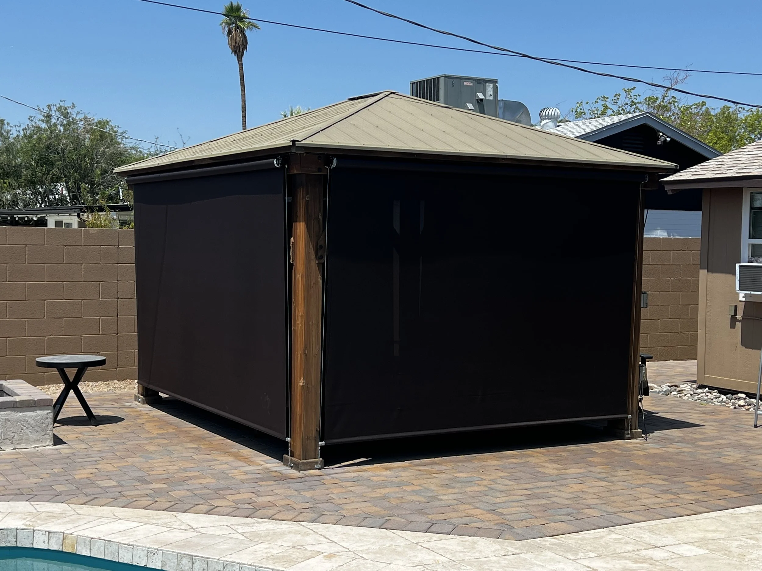 A black outdoor gazebo with a beige metal roof situated on a brick and stone paved backyard area, against a tan brick house wall with an air conditioning unit, and a small black side table nearby.