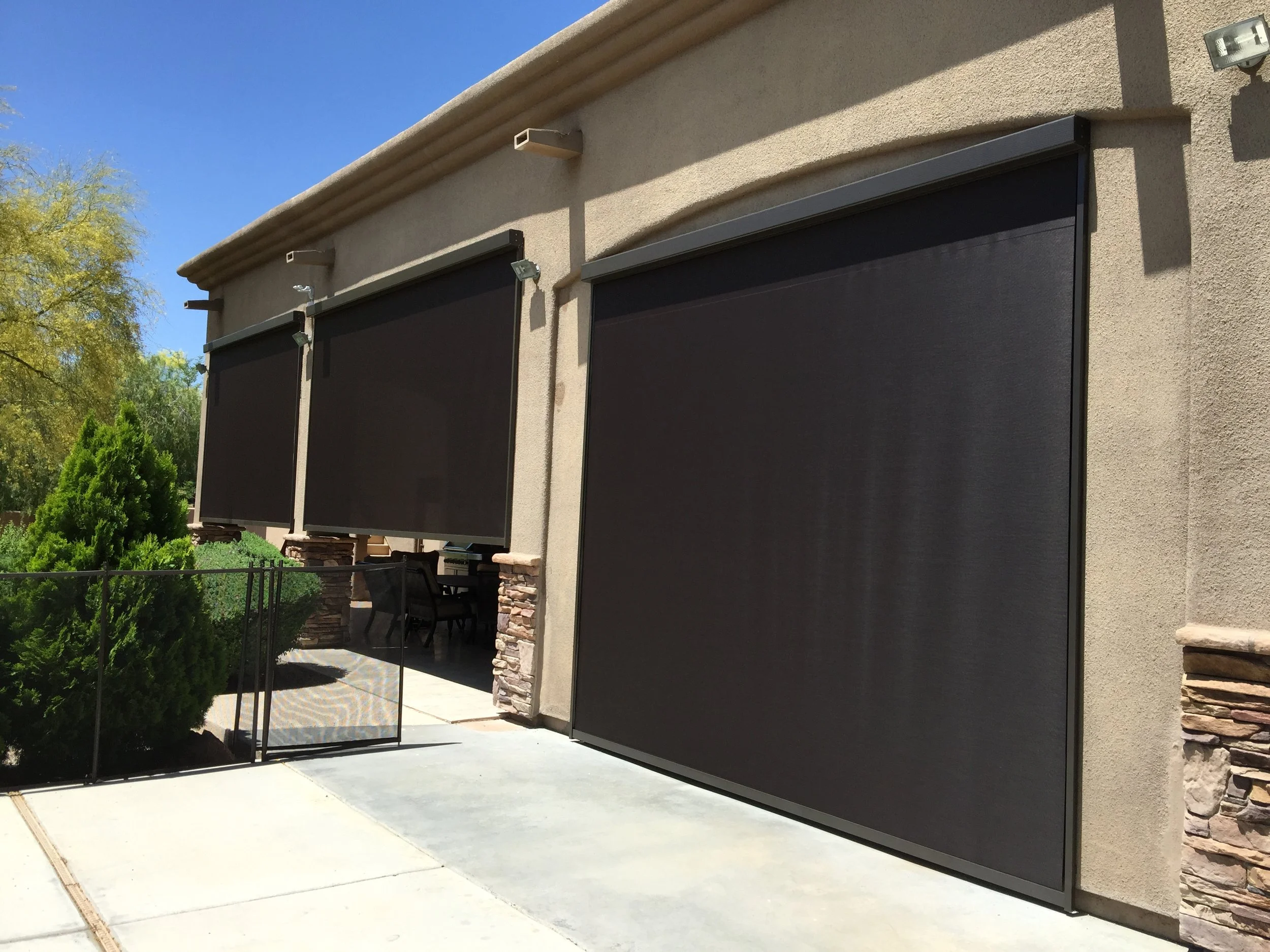 Three large black retractable garage screens on a beige stucco house, with a small safety gate and green bushes in front, sunny day with blue sky.