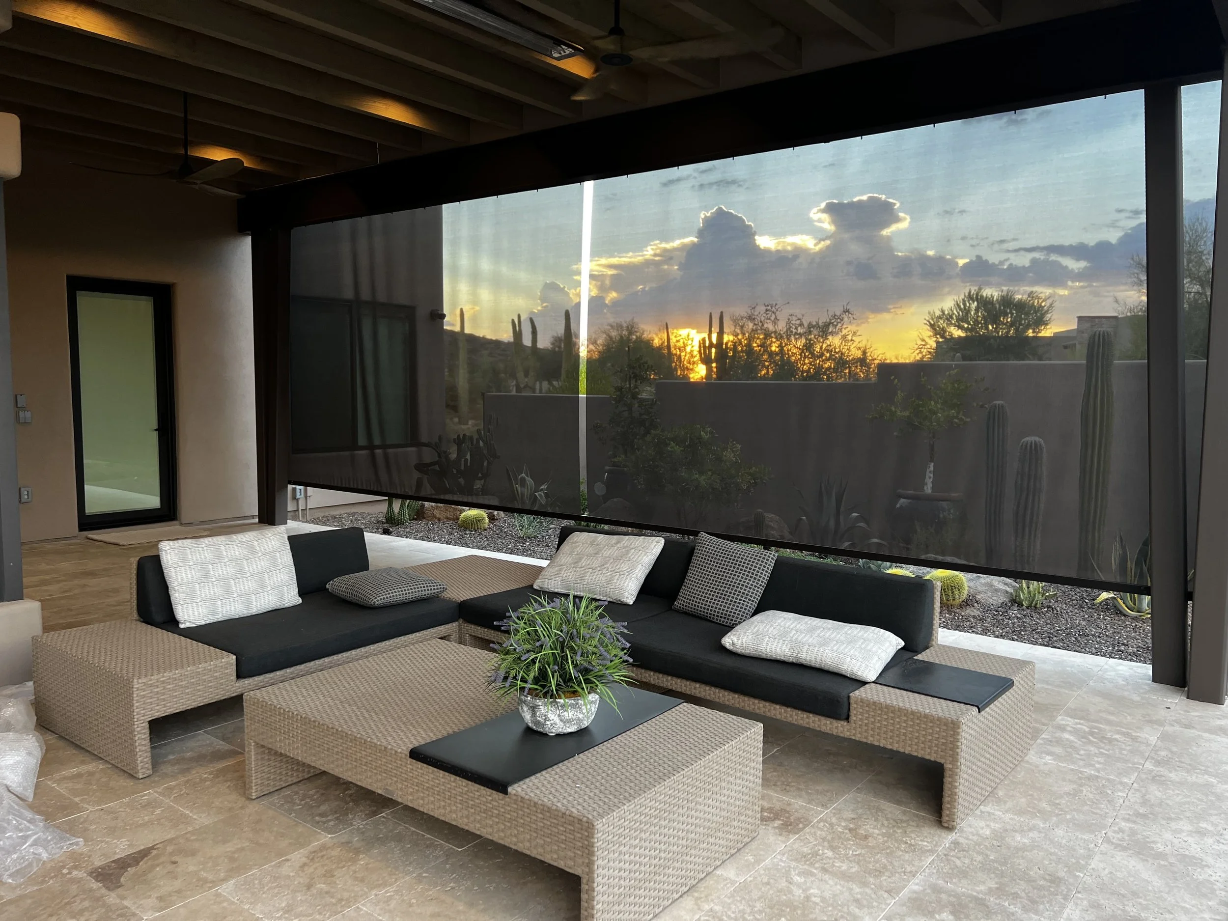 Living room with woven sectional sofa, plant centerpiece, and large screened window showing desert landscape with cacti and a sunset sky.