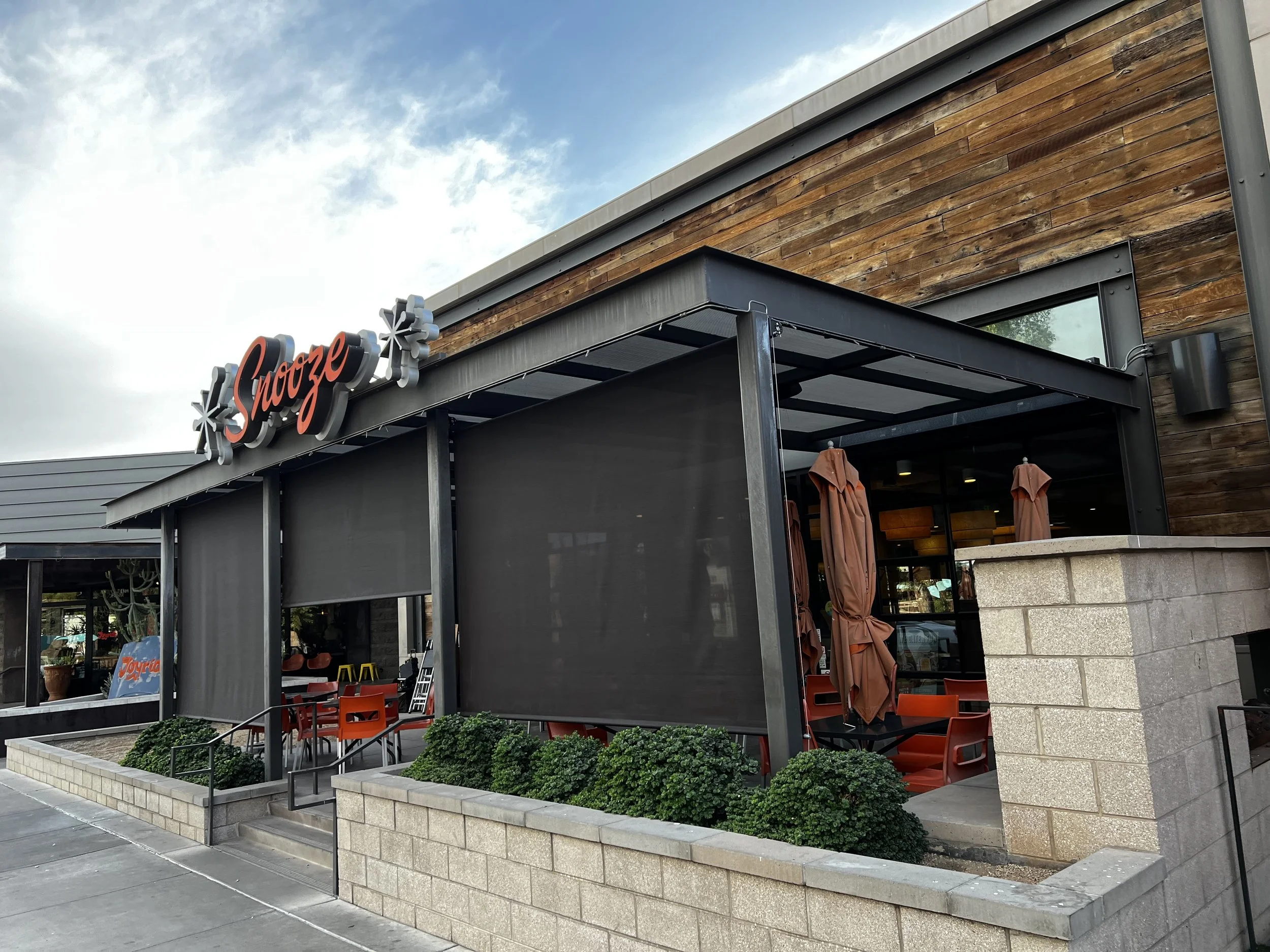 Exterior view of a restaurant called Spooze with a black awning, outdoor seating, and closed brown umbrellas. The building has wooden and brick exterior walls, and a sign with the restaurant's name in red and black letters.