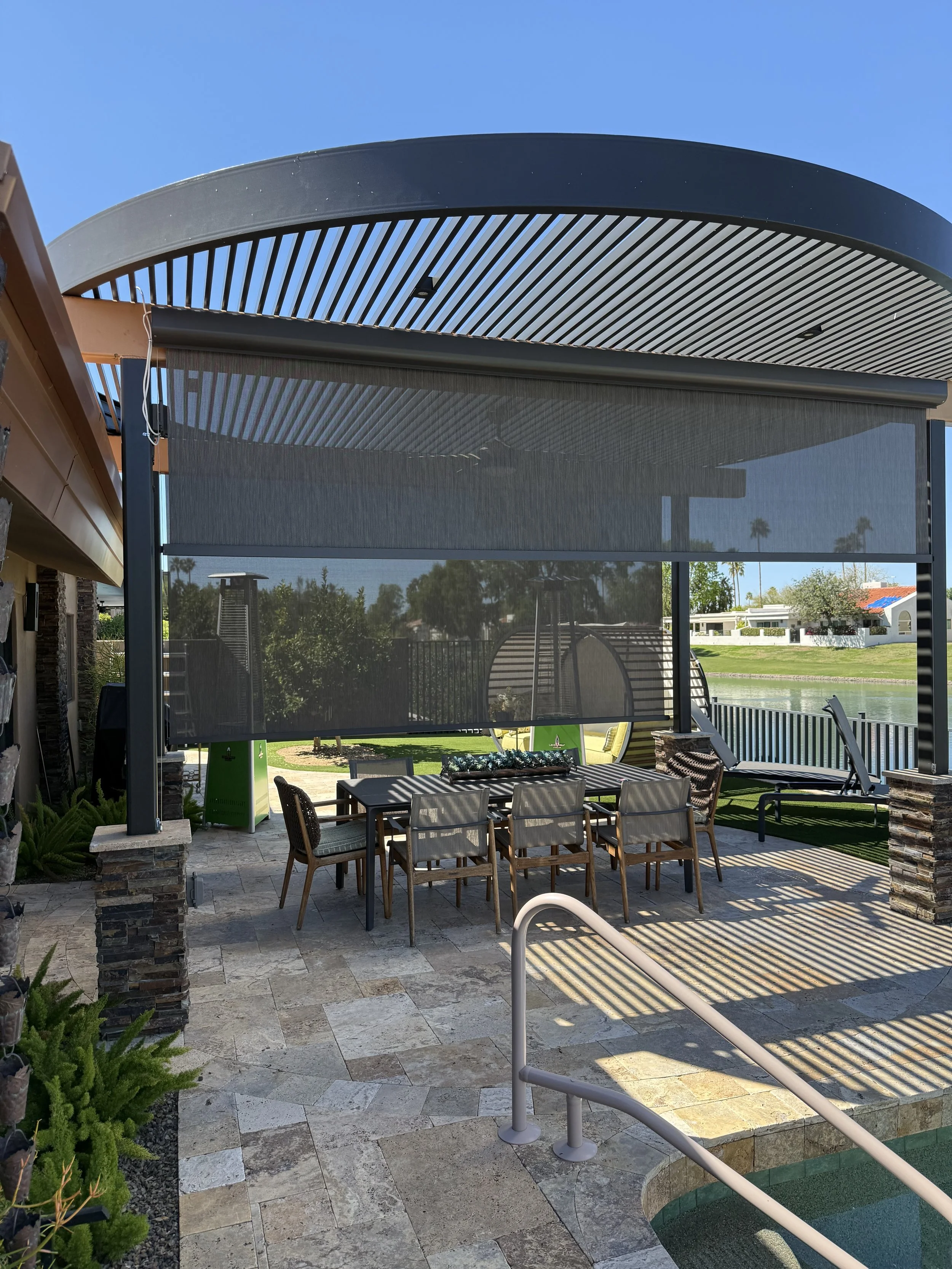 Outdoor patio area with a dining table and chairs underneath a black pergola, overlooking a lake with houses and trees in the background.