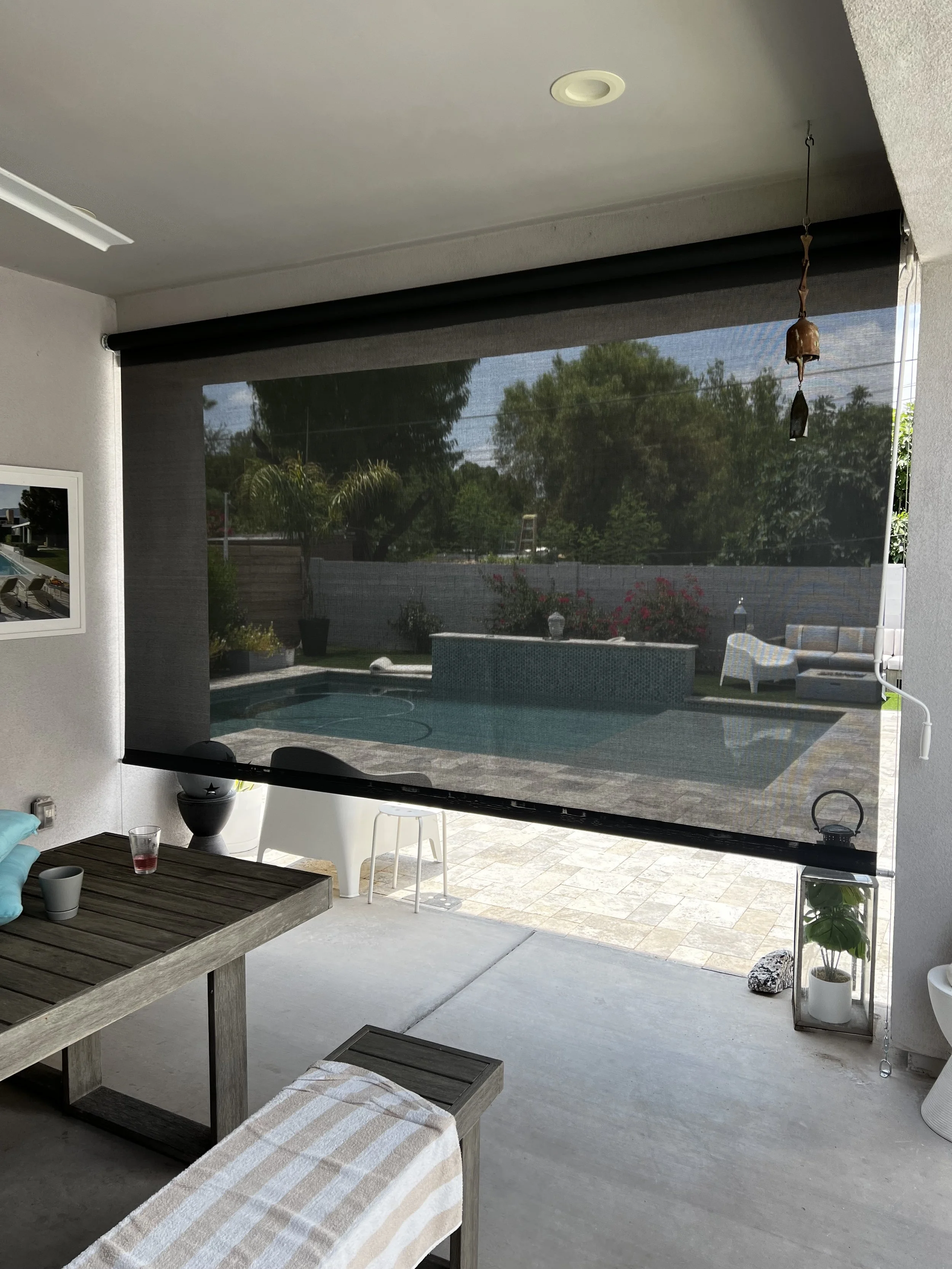 Indoor patio with view of backyard pool area, outdoor seating, potted plants, and trees beyond the fence, via a rolled-down screen.