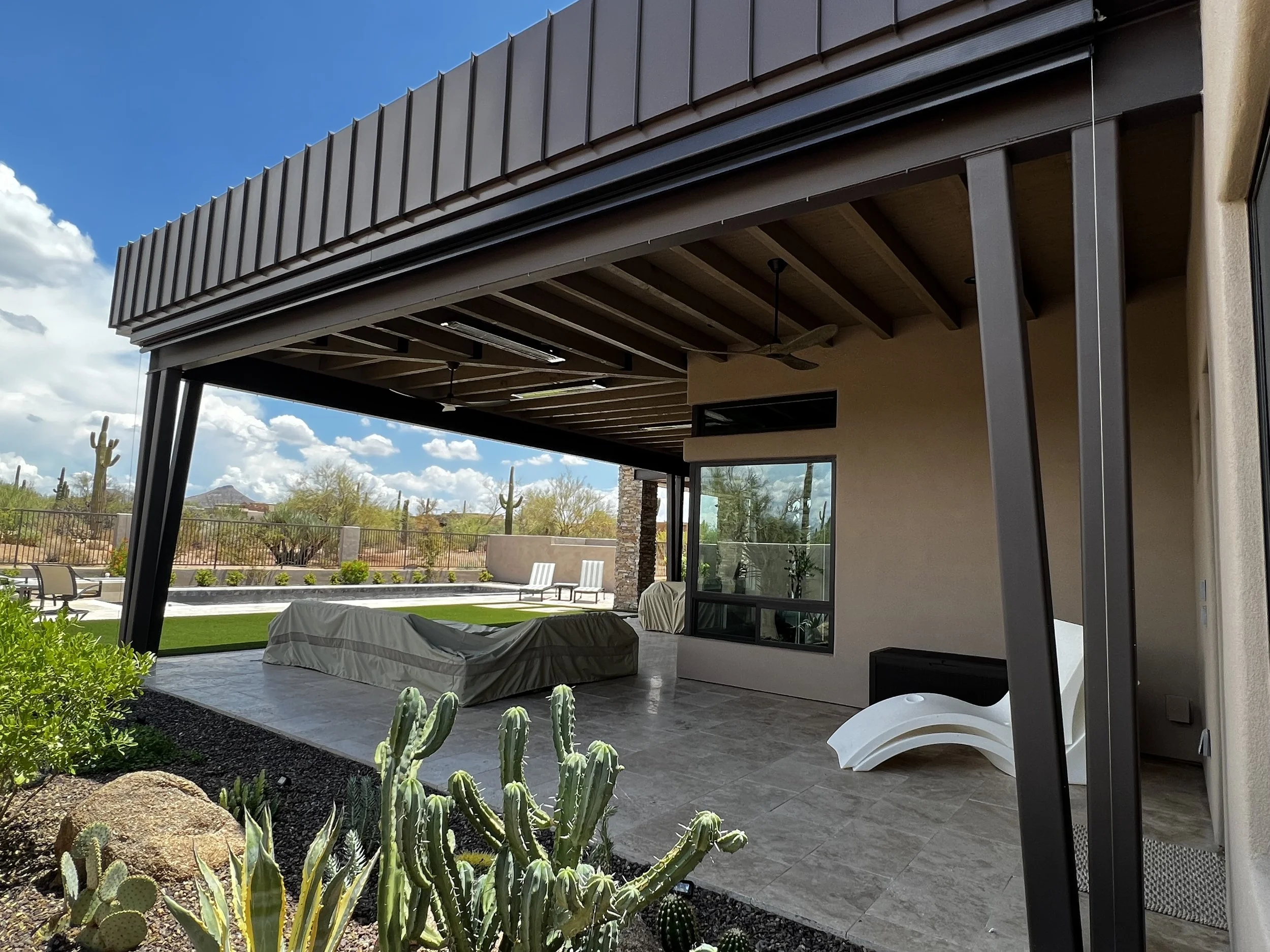 Covered patio area with outdoor furniture, potted plants, and desert landscaping including cacti, with a view of a backyard and desert landscape beyond, under a partly cloudy sky.