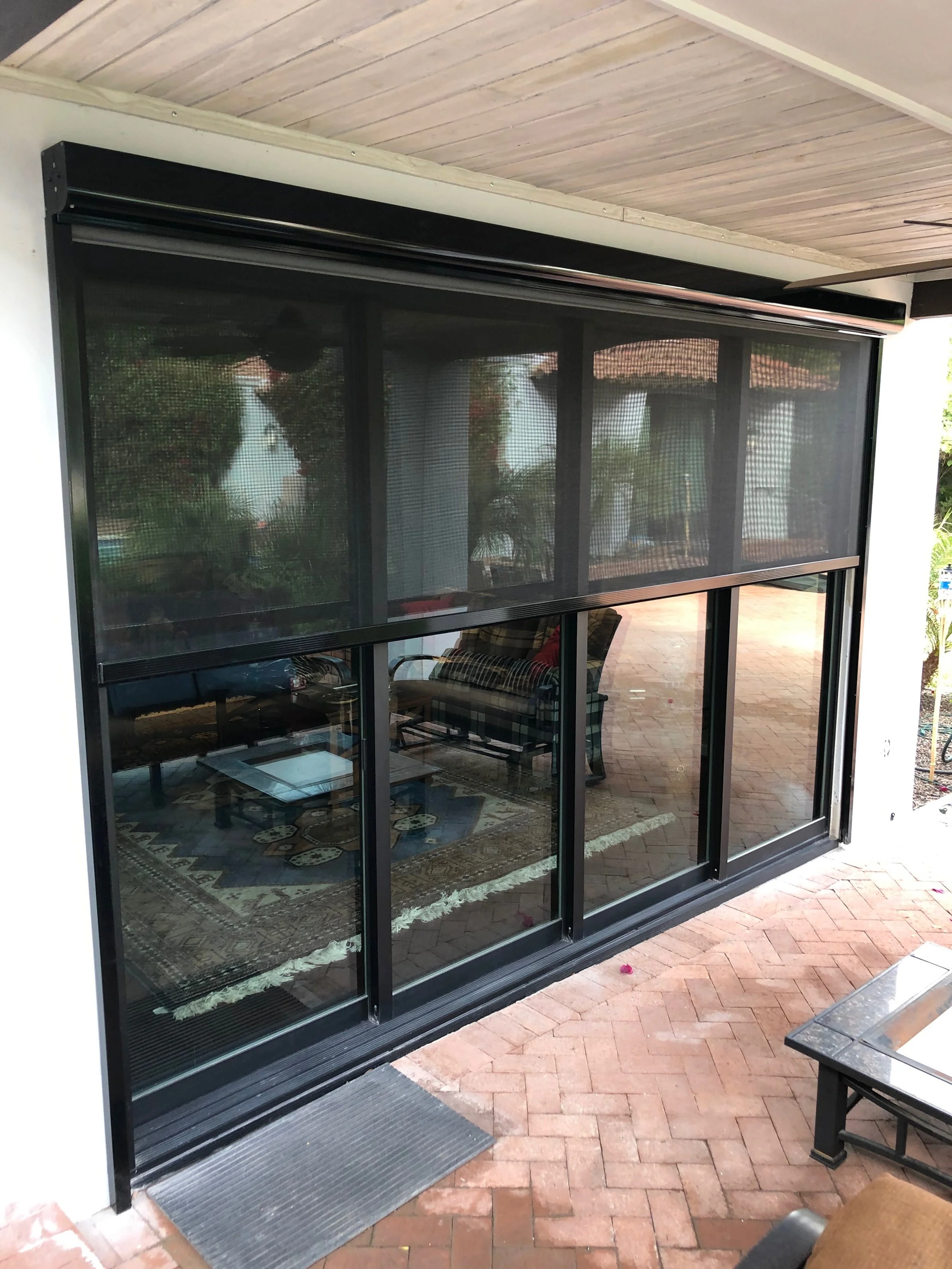 Sliding glass door with a screen, leading to a patio area with outdoor furniture and a patterned rug, brick paving, and trees in the reflection.