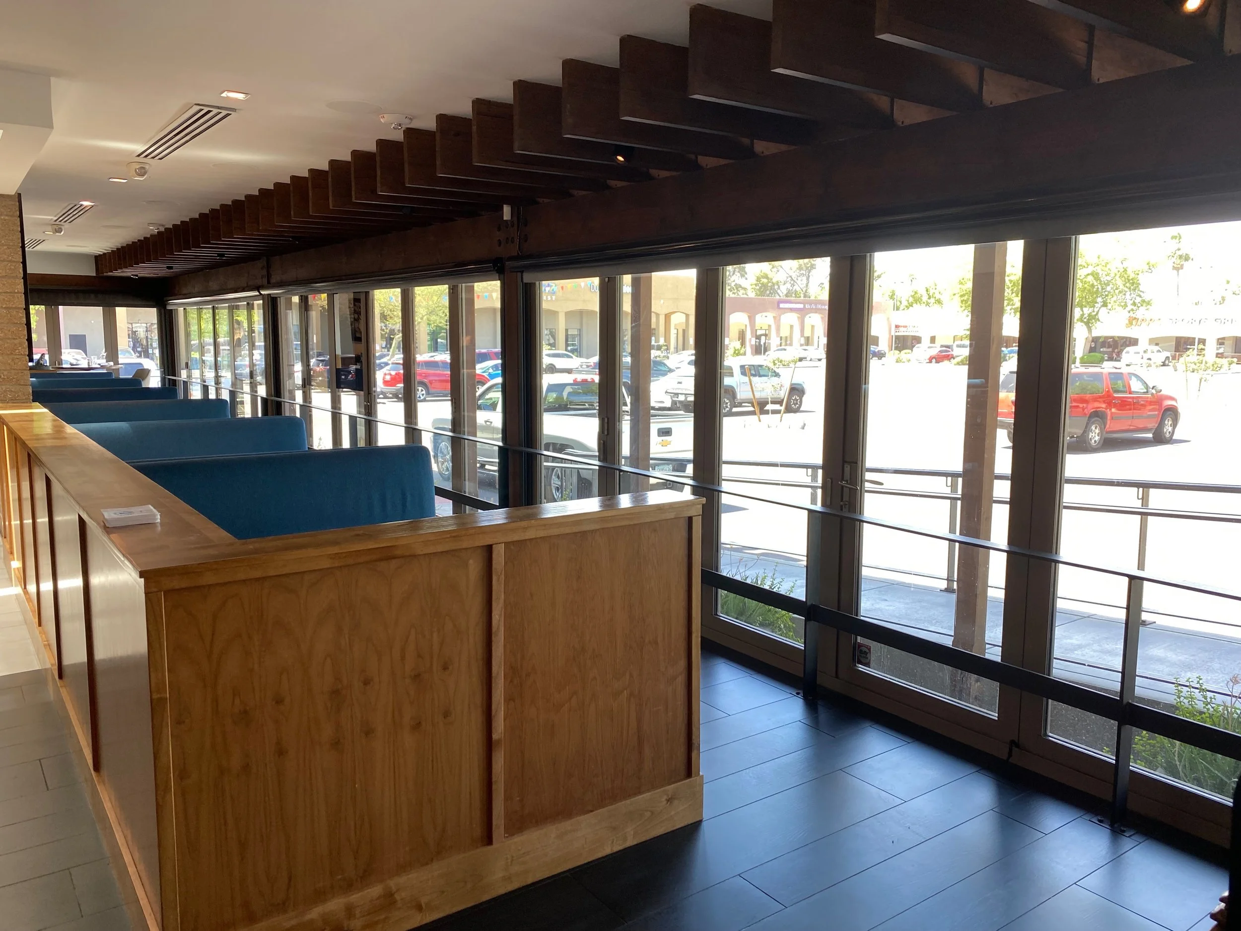 Interior of a restaurant or cafe with blue booths along a wall of large glass windows showing parked cars outside, wooden accents, and a ceiling with horizontal wooden slats.