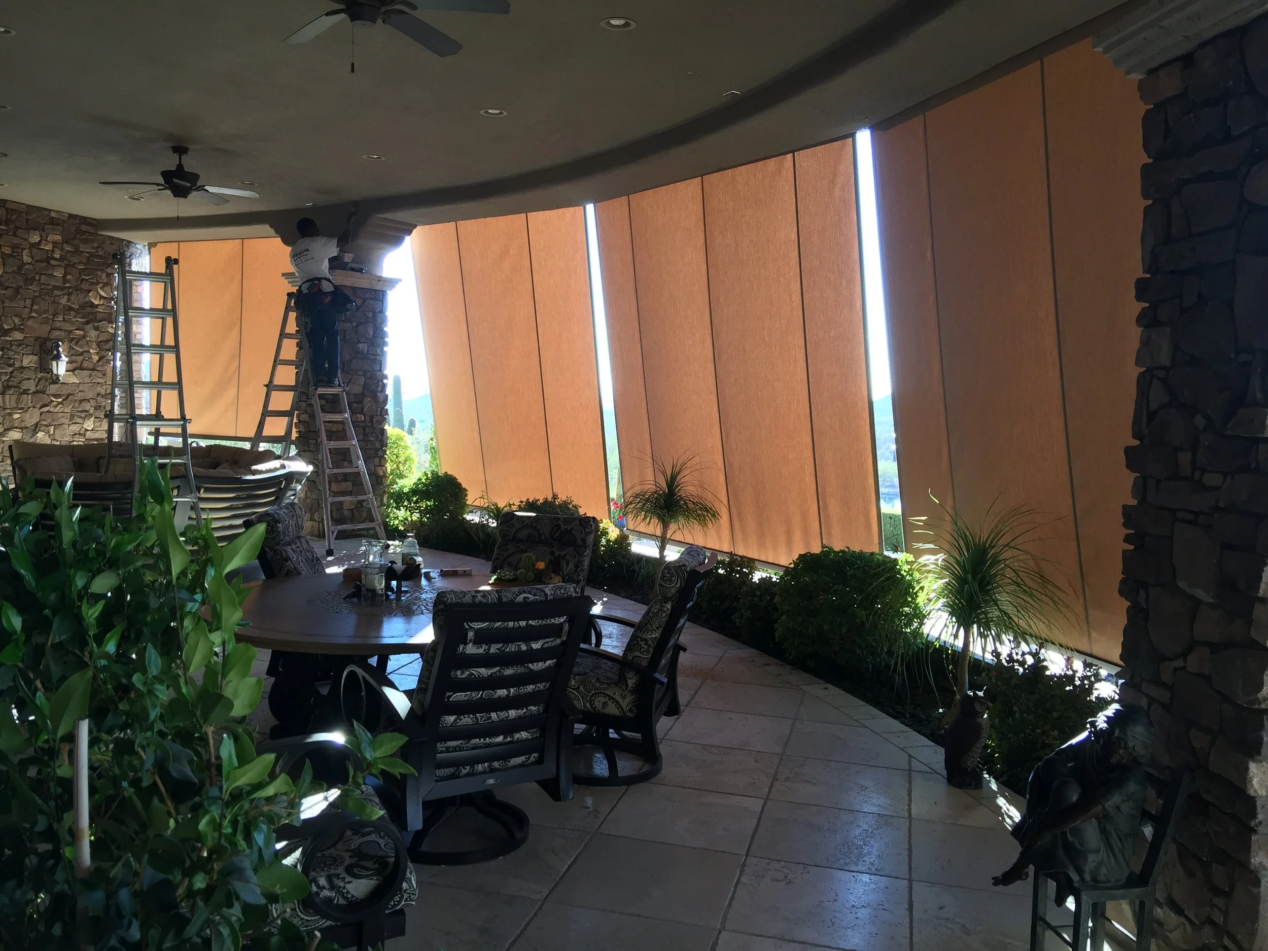 Indoor patio with a round table and chairs, potted plants, stone pillars, and large brown window coverings, with two workers on ladders adjusting the window coverings.