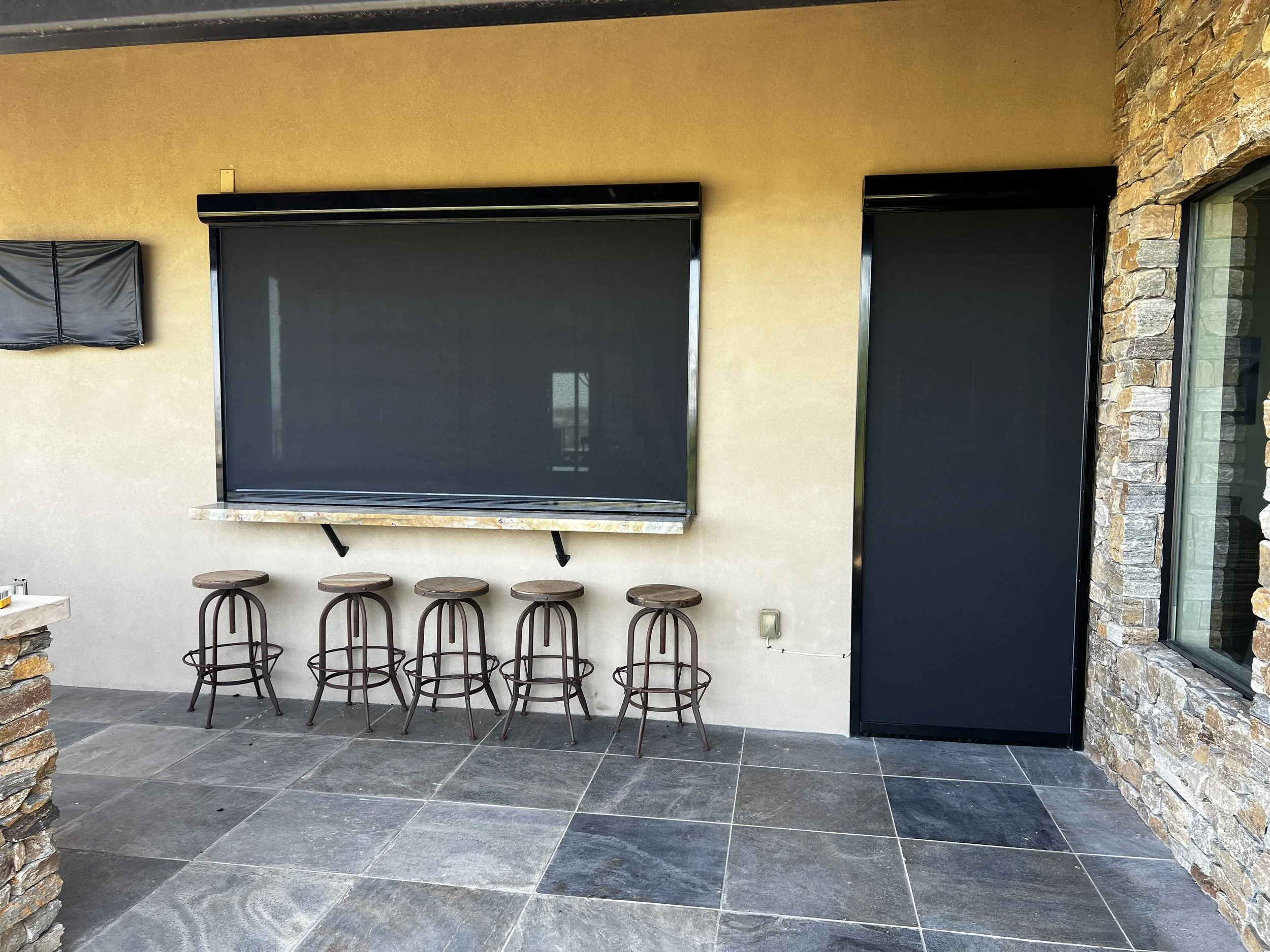 Exterior patio with six metal bar stools, a stone countertop, and two black roll-up patio shades on a beige stucco wall with stone accents.