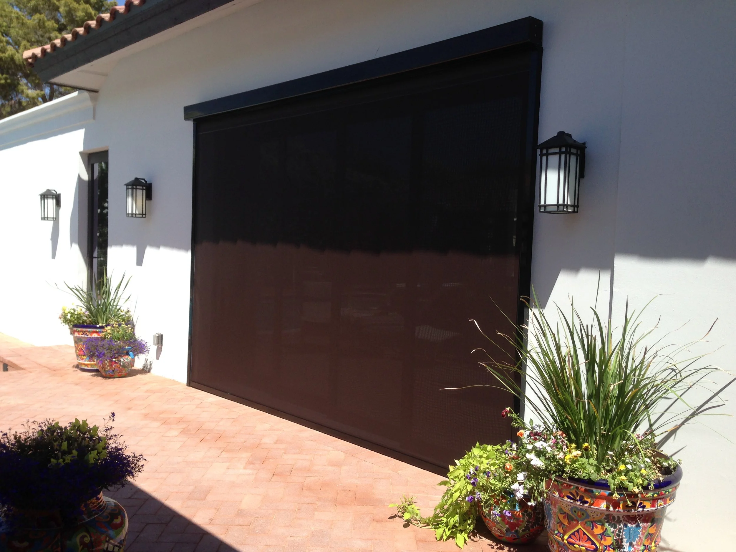 Exterior of a house with a large black garage door, white walls, and four wall-mounted black lanterns. Potted plants with colorful decorative pots and green foliage are on a brick patio.