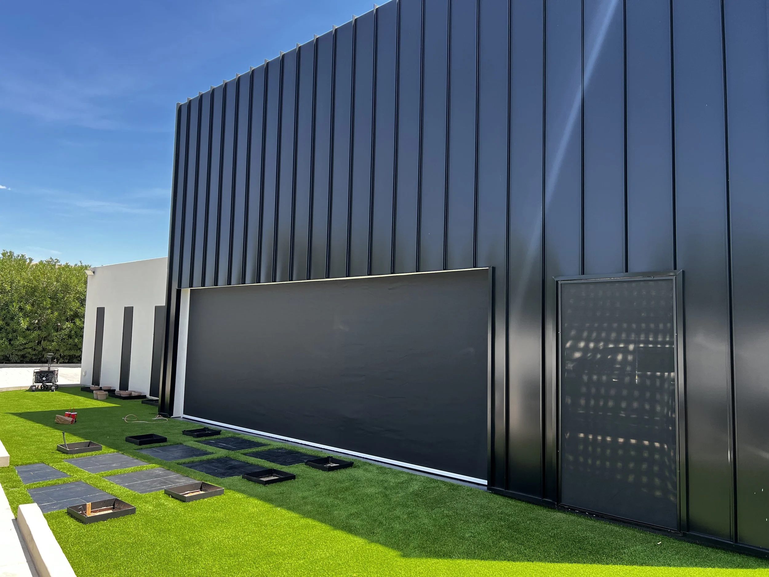 Modern black exterior building with vertical metal panels, small window, and a green artificial grass area with square black landscape tiles, some construction materials and tools.