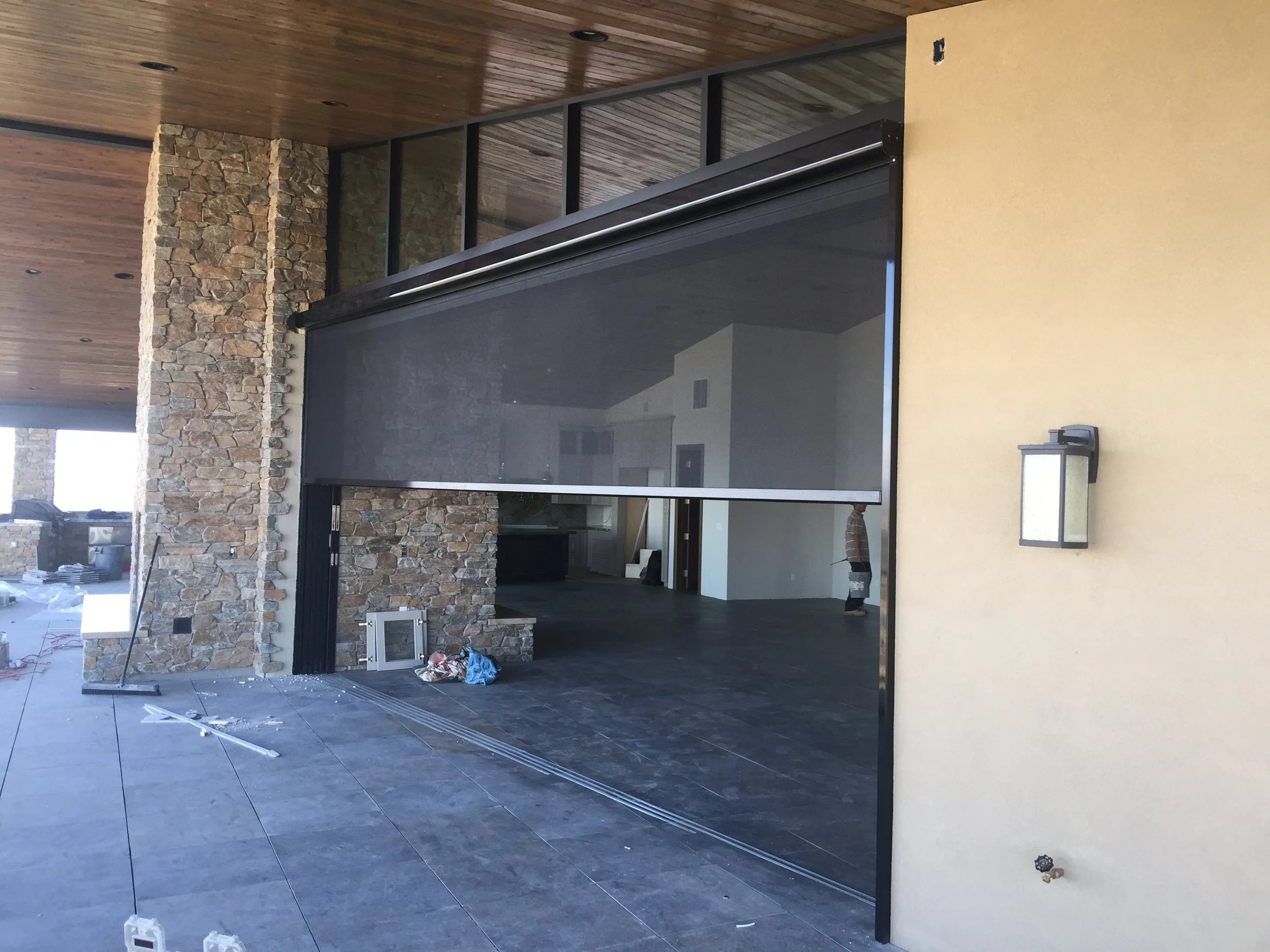 Modern outdoor patio area with sliding glass door, stone wall, and beige wall with wall-mounted light fixture under a wooden ceiling, with construction materials on the floor.