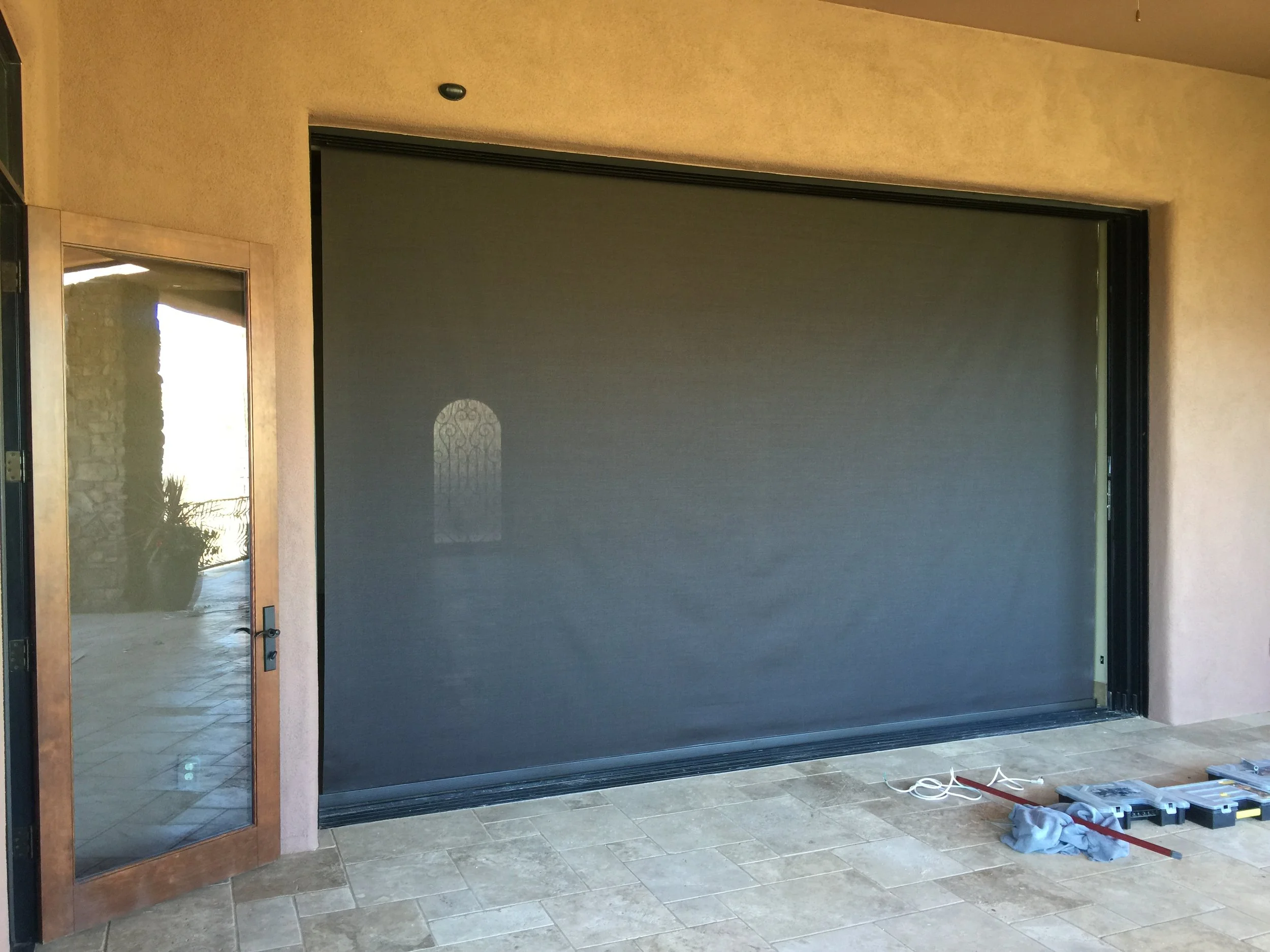 A large black retractable security screen door installed on a patio with a wooden framed glass door on the left. Tools and materials are on the floor in front of the door.