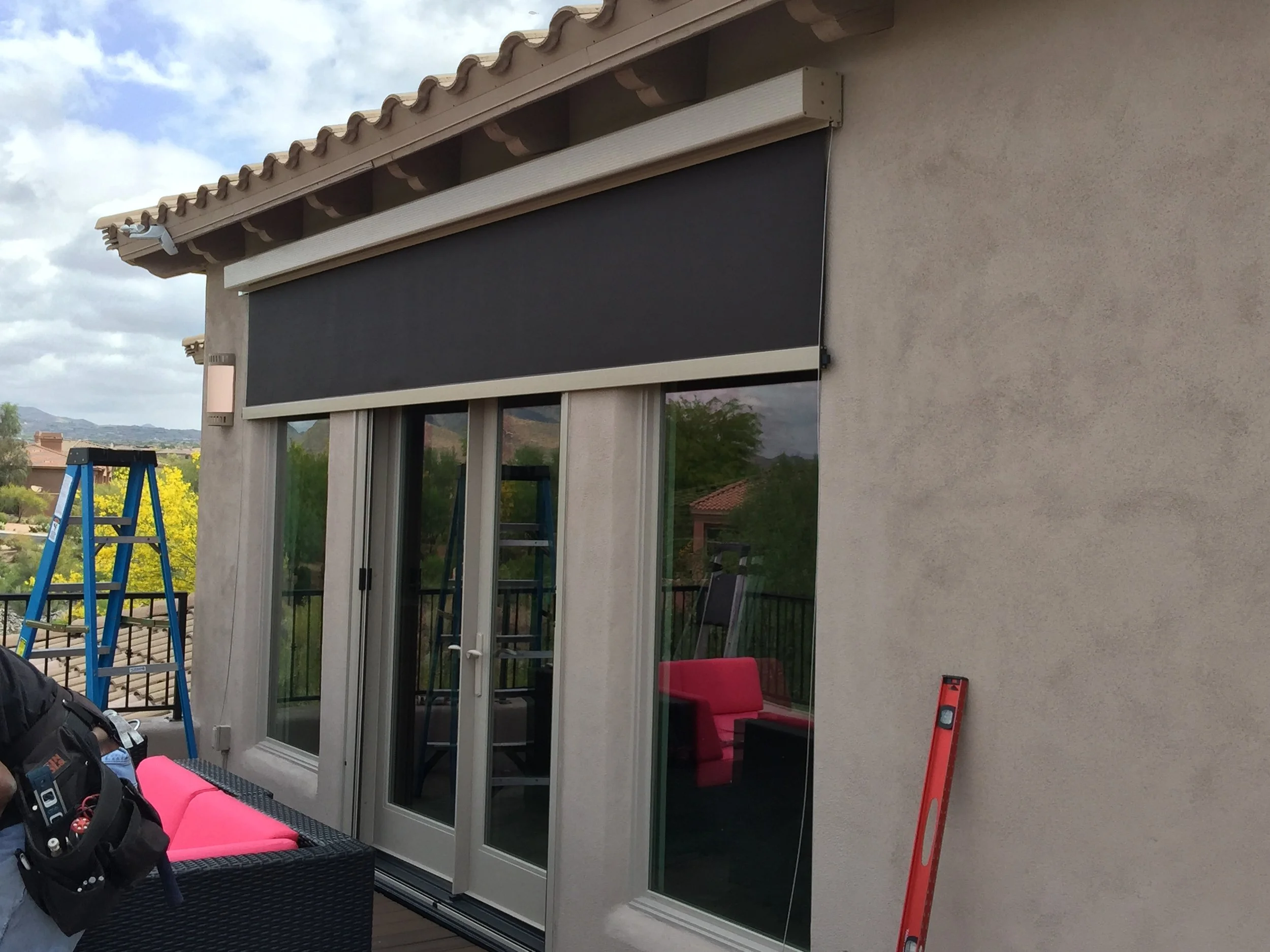 Backyard patio with sliding glass door, black retractable awning, pink outdoor seating, blue ladder, and a level leaning against the house wall.