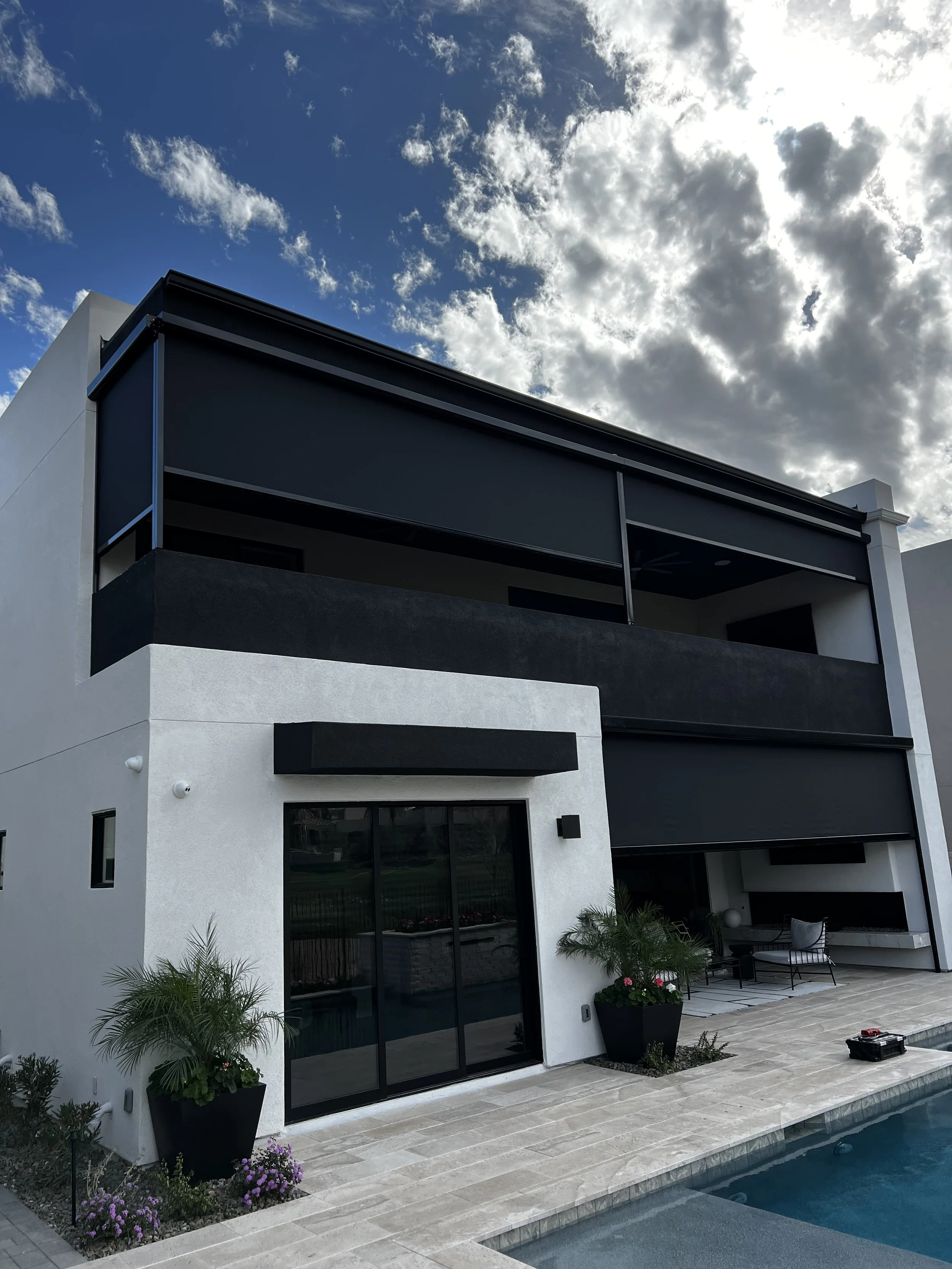 Modern white and black multi-story house with a balcony, patio furniture, potted plants, and a swimming pool in the backyard under a partly cloudy sky.