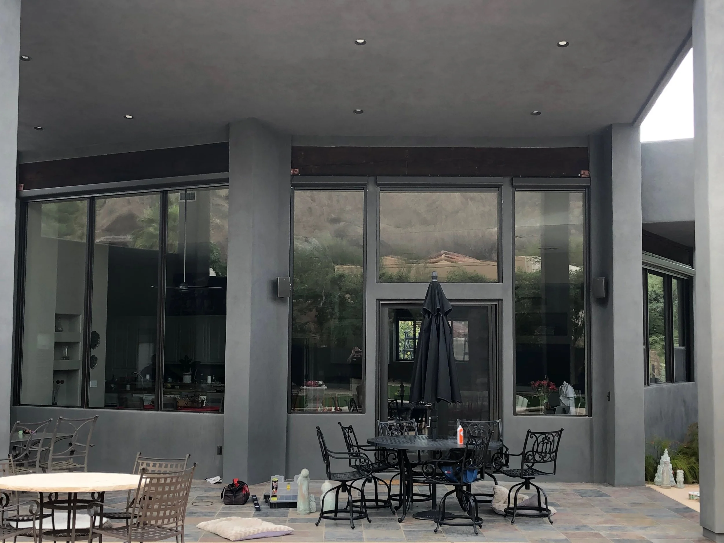 An outdoor patio area in front of a modern house with large glass windows and sliding doors, featuring a round table with chairs, a closed black umbrella, and some miscellaneous objects on the patio floor.
