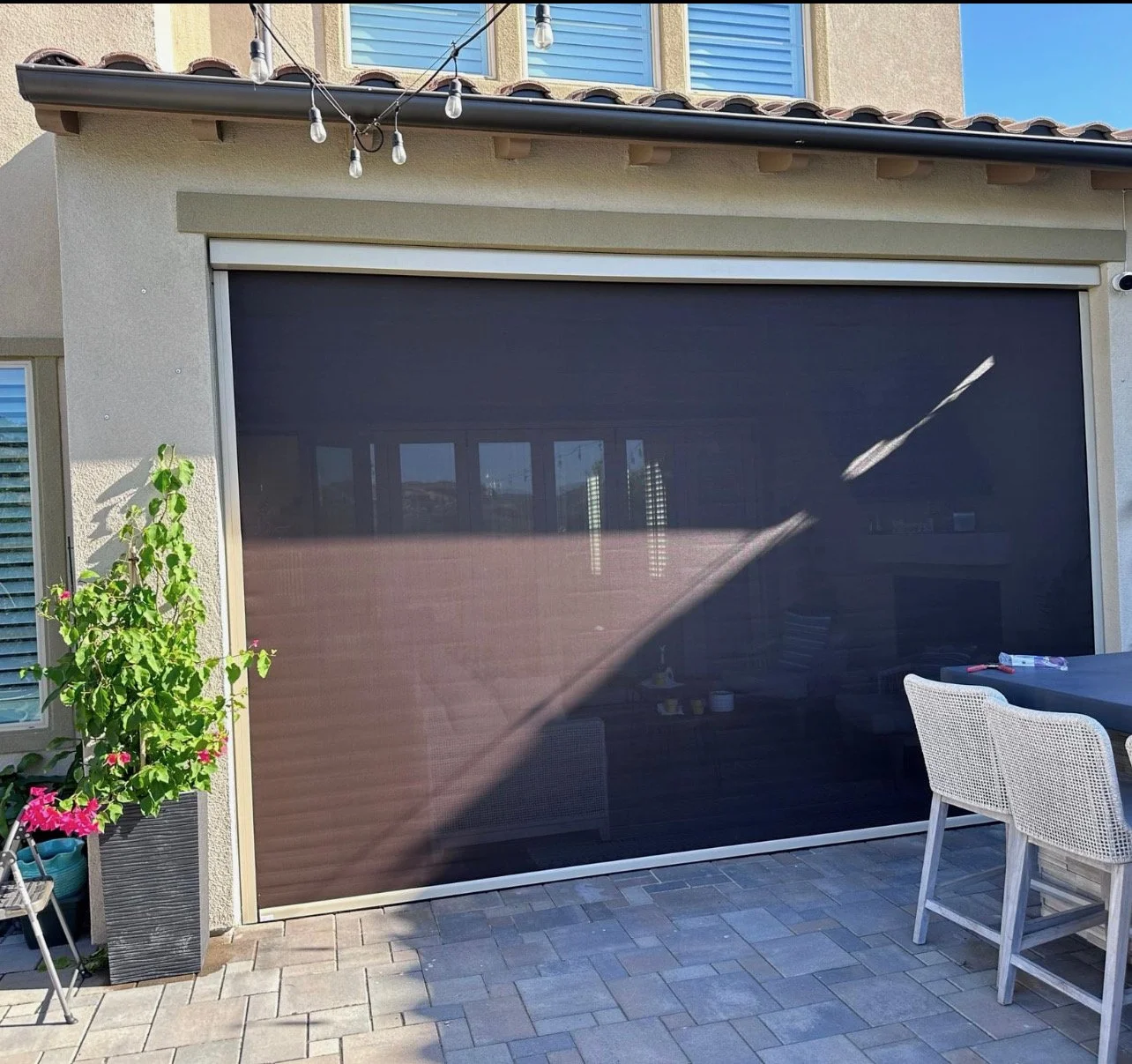 A backyard patio with a large black retractable screen door, a potted plant with pink flowers on the left, and a white outdoor dining table with white rattan chairs on the right. String lights hang above.