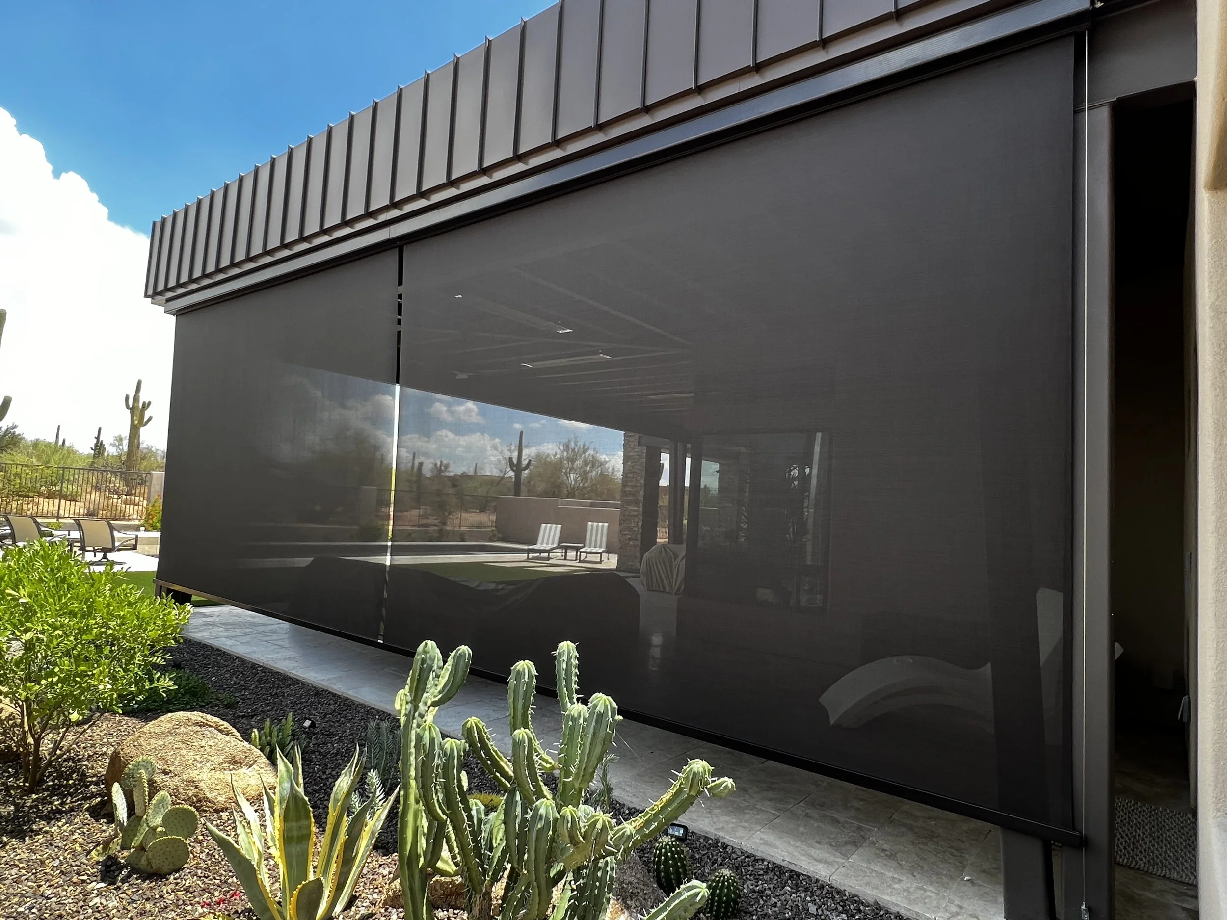 Modern house with a large black shade screen, desert plants in the foreground, and blue sky with clouds in the background.