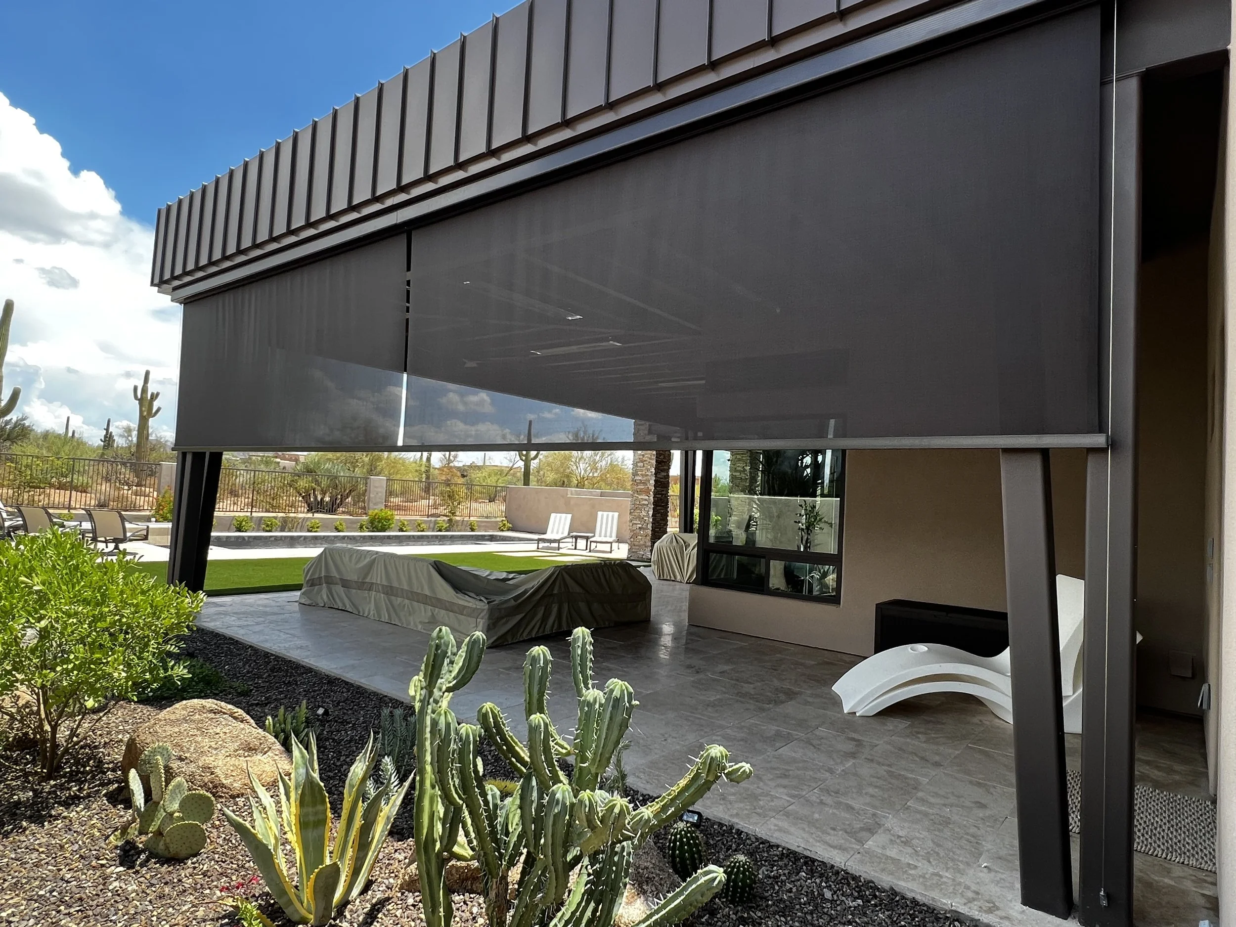 Modern outdoor patio area with a covered sitting space, cactus plants in the foreground, and desert landscape with saguaro cacti in the background.