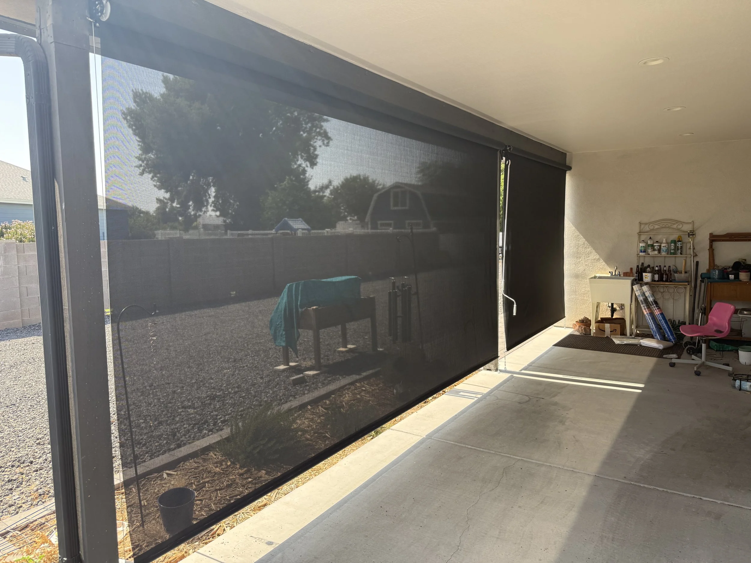 A covered patio with a black retractable screen door, a small garden area with gravel, a garden bench with a green towel, and assorted outdoor and gardening supplies, with sunlight casting shadows.