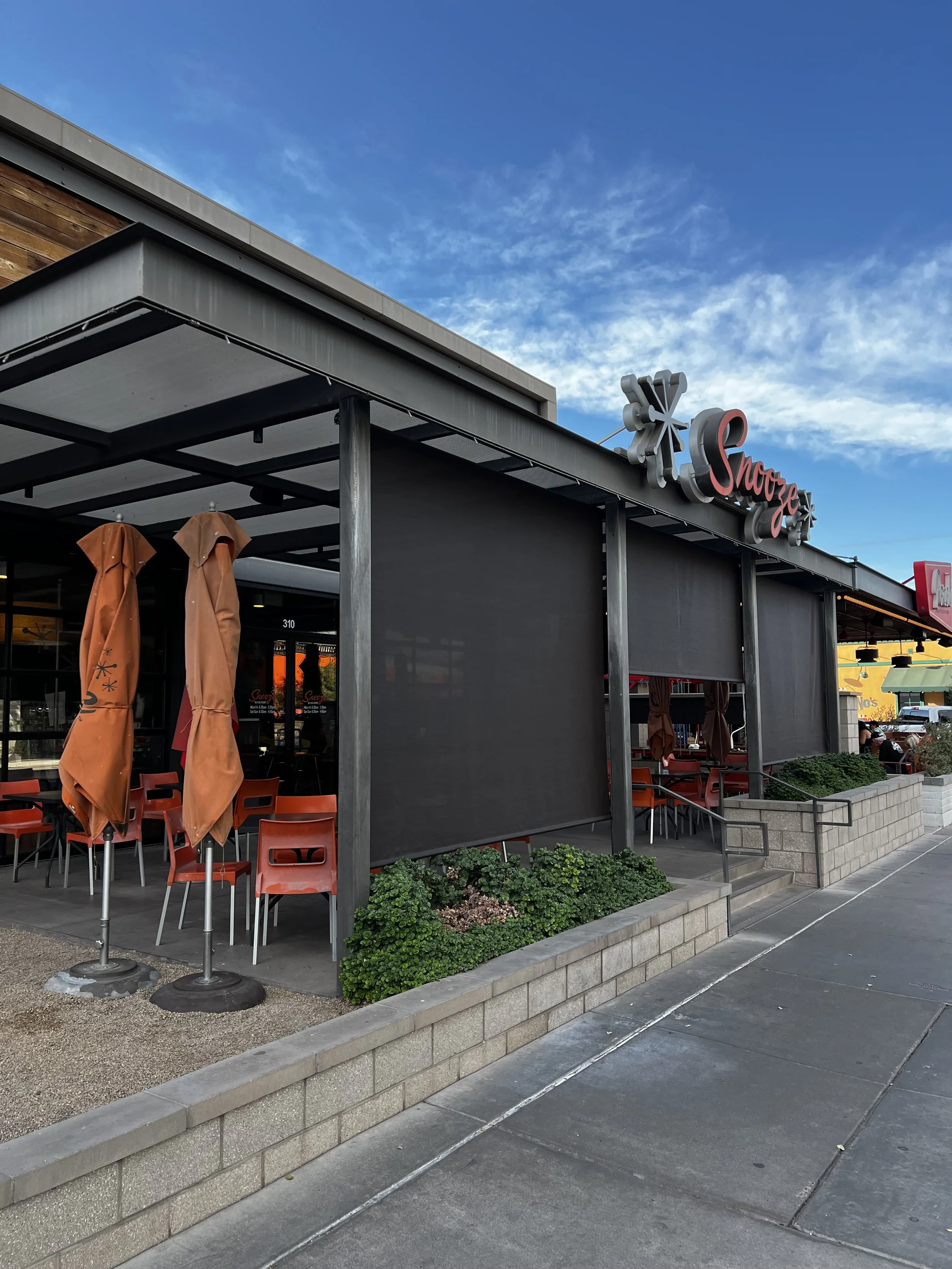 Outdoor patio of a restaurant named Snooze with orange chairs, closed orange umbrellas, and black retractable shades under a partly cloudy sky.