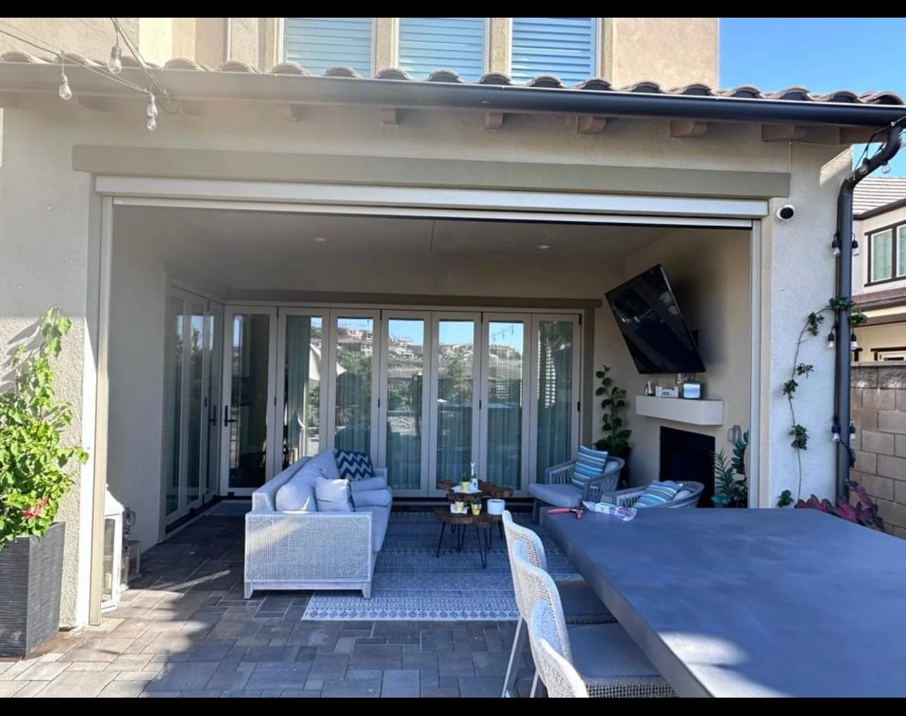Outdoor patio area with white seating, striped cushions, a rug, and large glass sliding doors leading inside. There is a television mounted above a fireplace and string lights hanging from the roof. The space is decorated with plants and has a bright