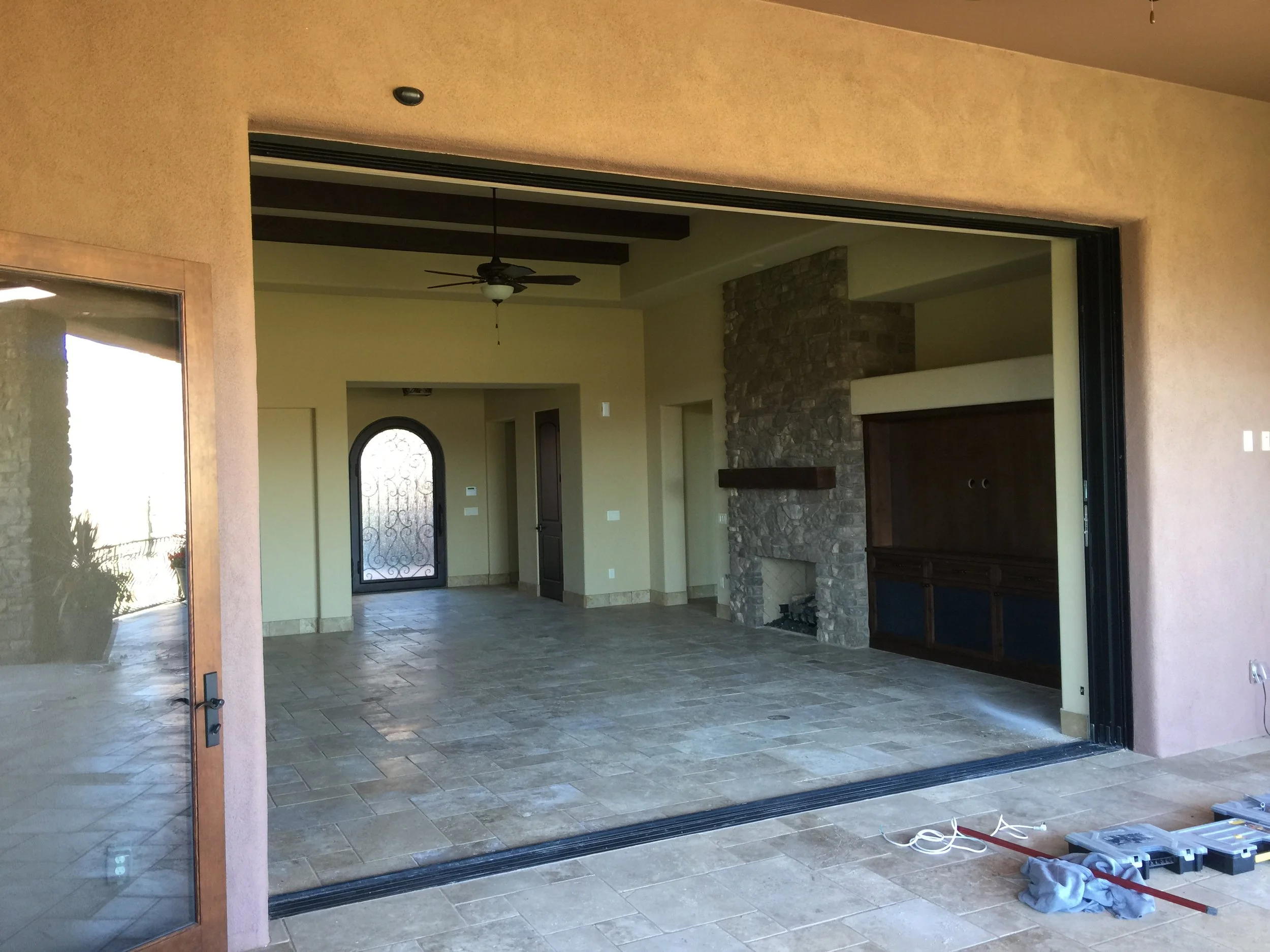 Empty living room with stone fireplace, ceiling fan, and sliding glass door leading outside, under construction with tools and materials on the floor.