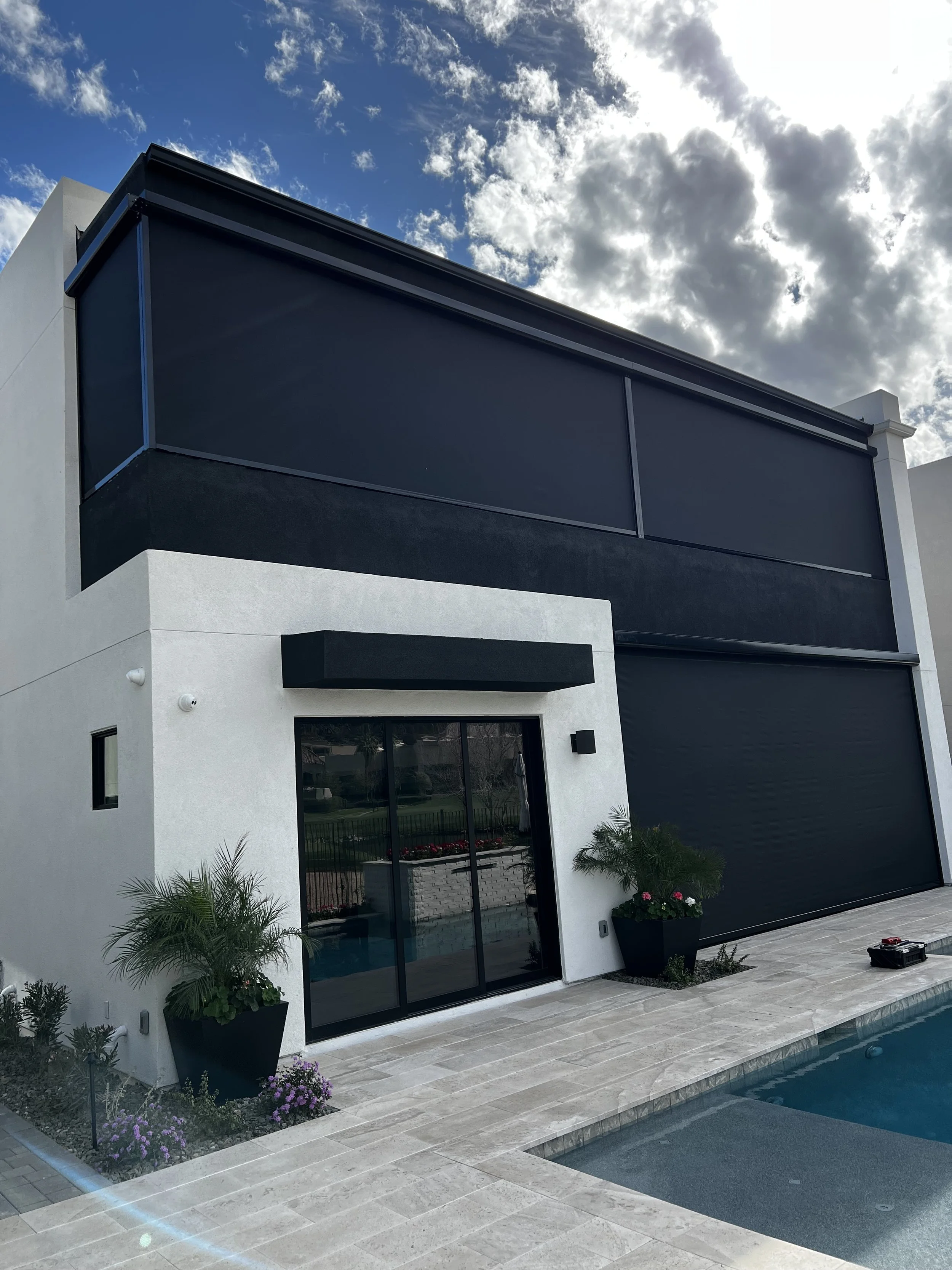 Modern two-story house with white exterior walls, black window shades, sliding glass door, and a black garage door, next to a swimming pool with potted plants and flowers, under a partly cloudy sky.