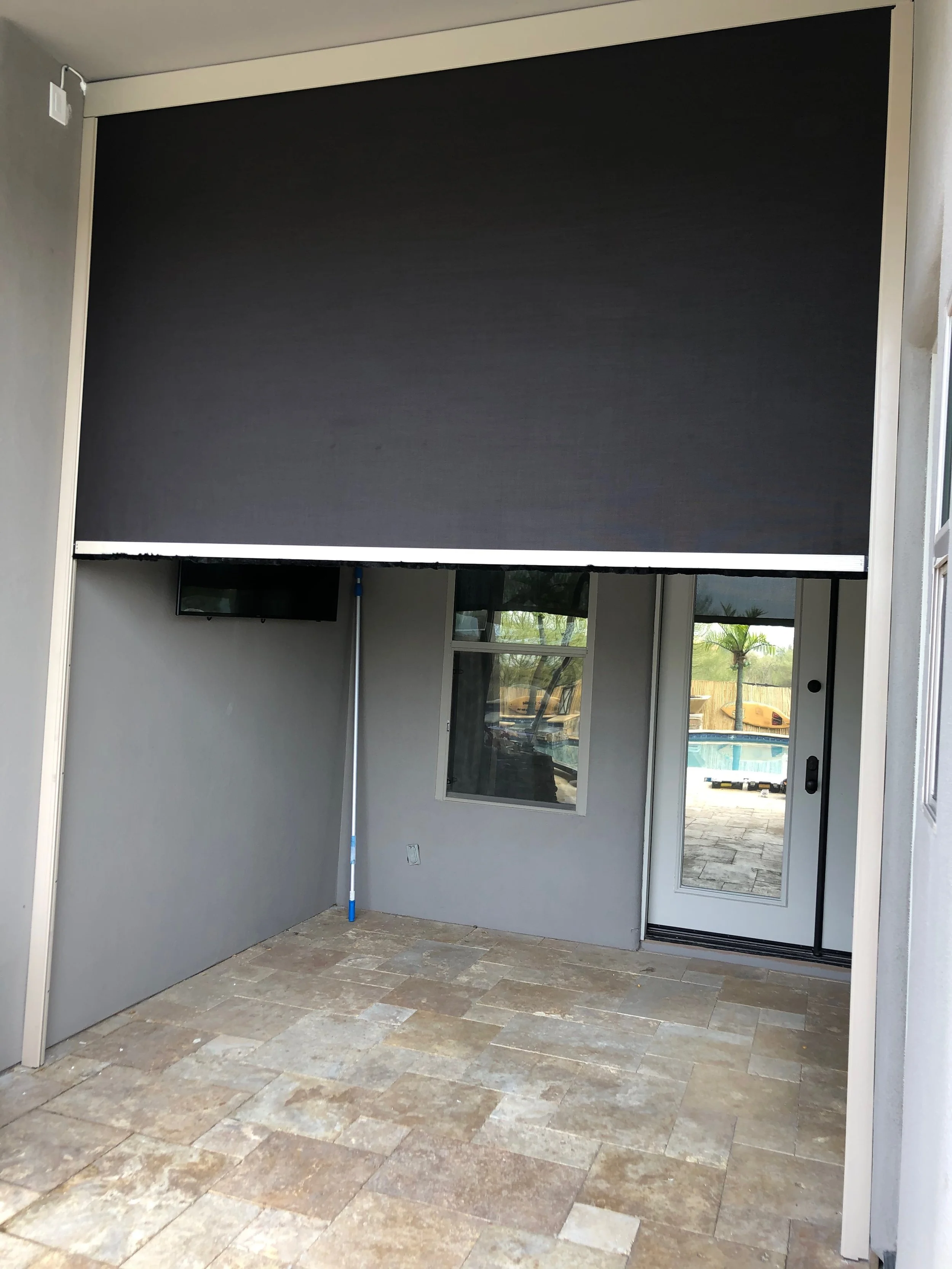 An outdoor patio area with stone tile flooring, a sliding glass door, two windows, and a large black retractable awning or shade covering the space.