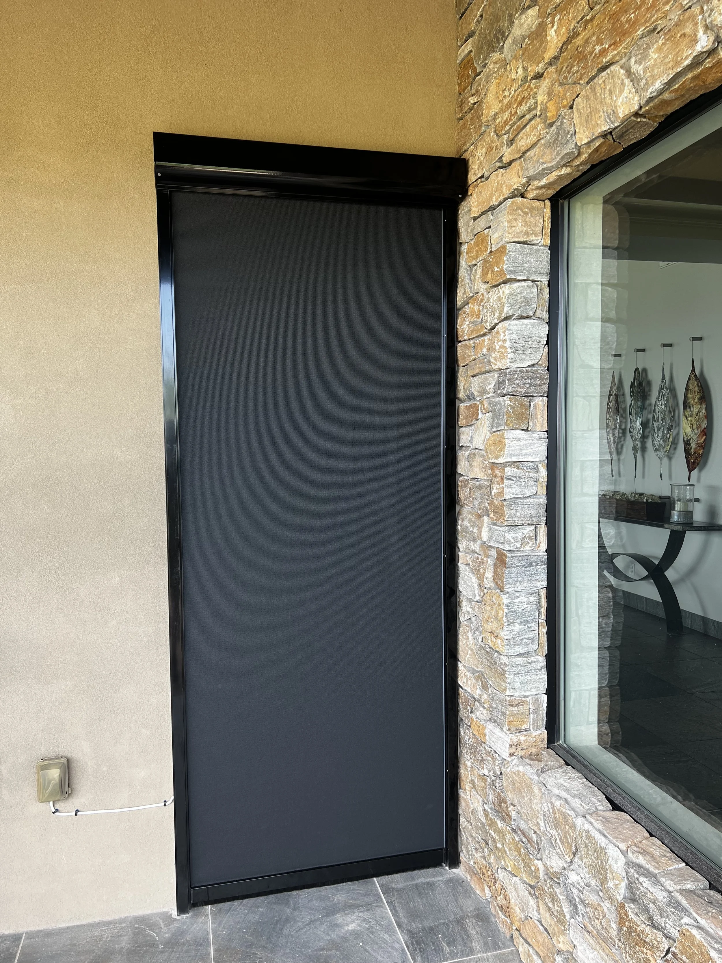 A black outdoor privacy screen or gate mounted next to a window with a stone frame, on a beige wall with a small electrical outlet.