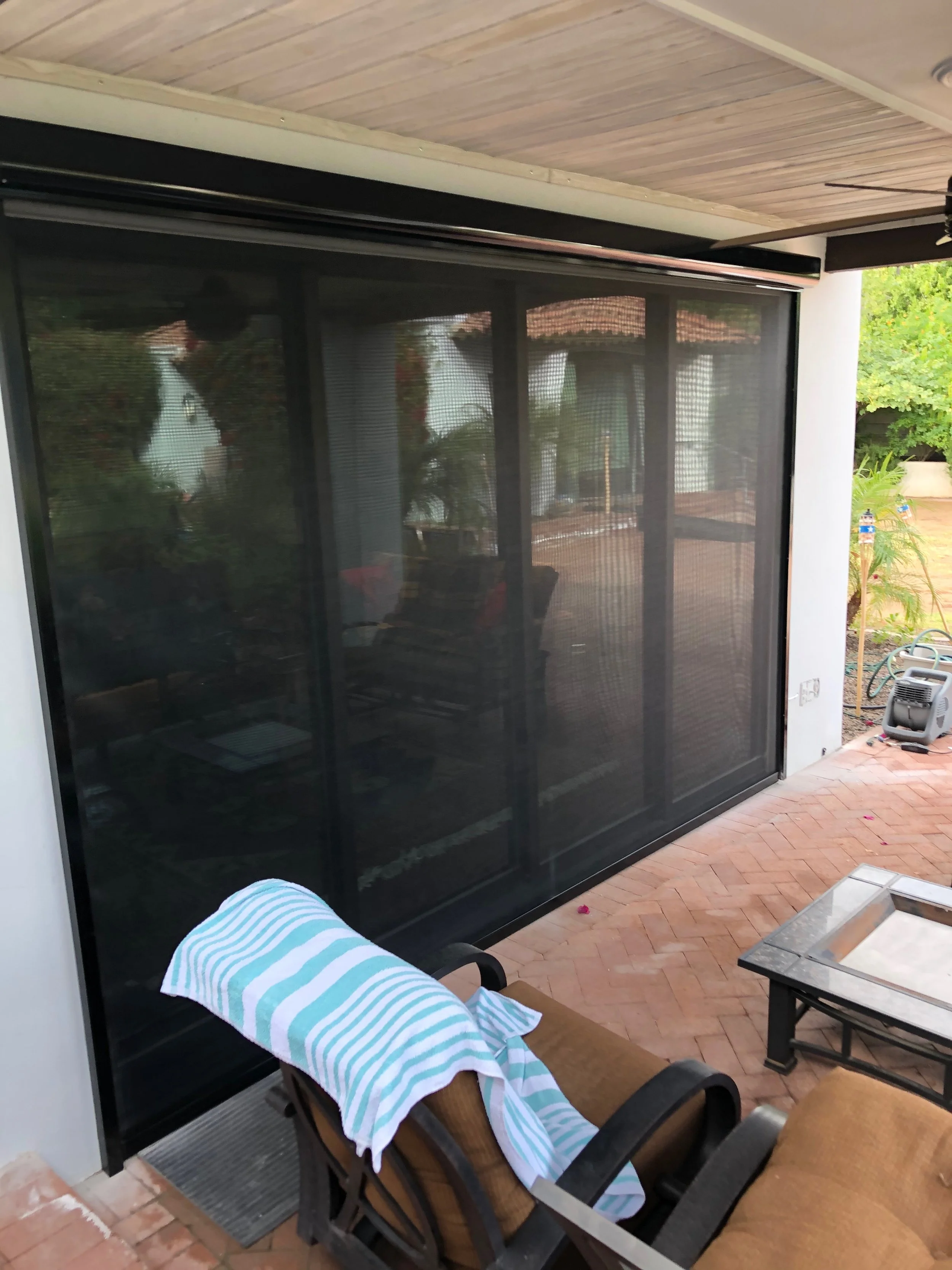 Close-up view of a patio door with a black mesh screen; a lounge chair with a striped towel draped over it on a brick patio.