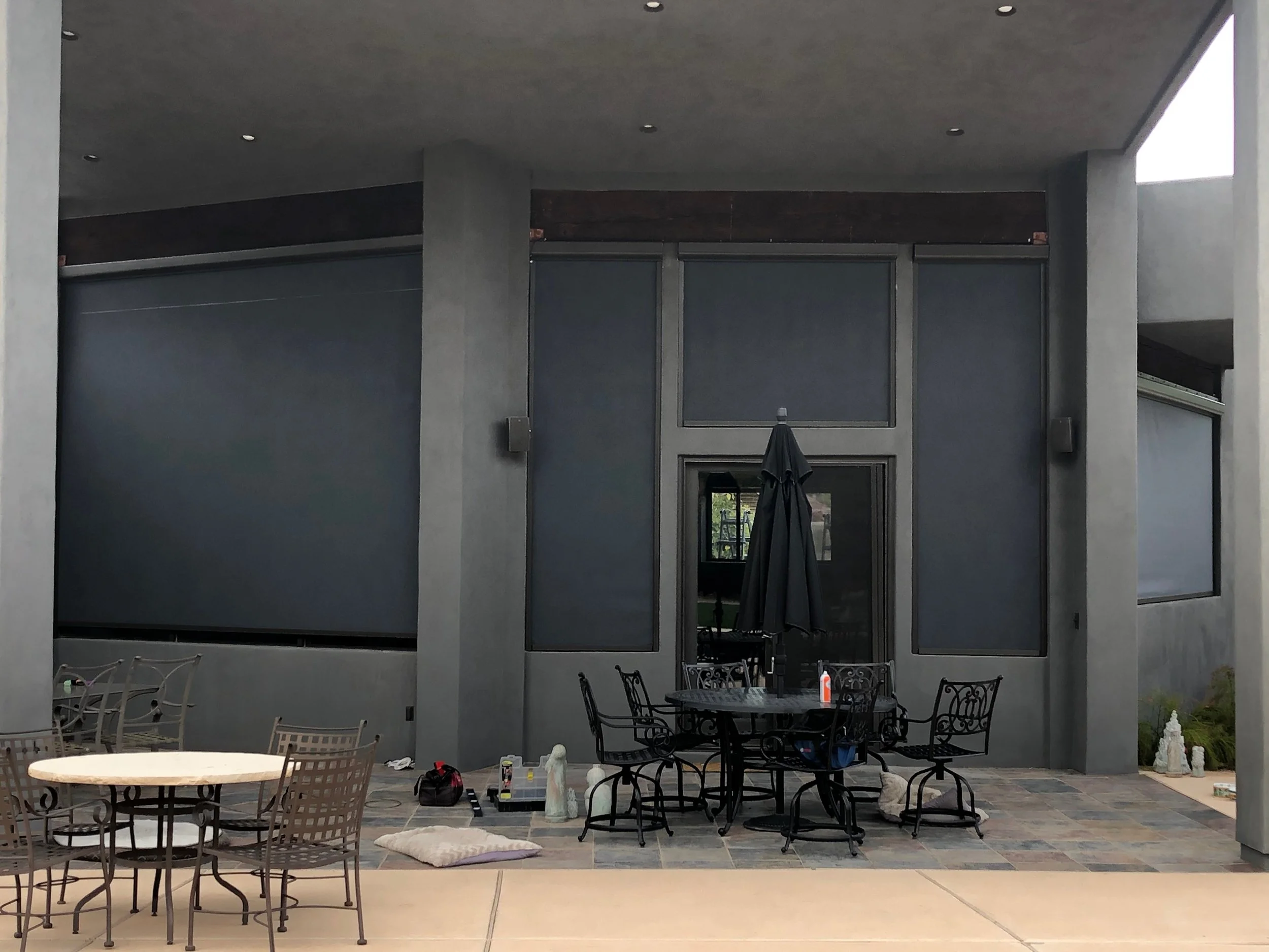 Outdoor patio with black metal table and chairs, umbrella, and various objects, with a dark gray wall and windows in the background.