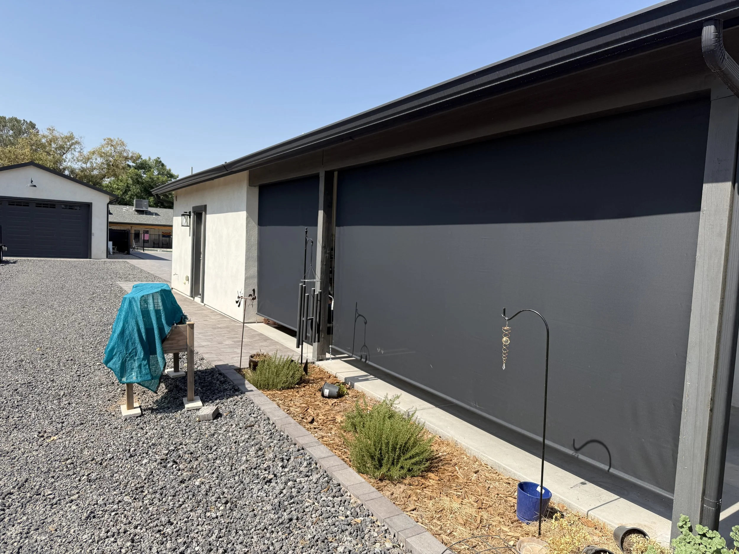 Exterior of a building with two dark gray garage doors, a concrete walkway, sparse landscaping with small plants, a blue bucket, and gardening tools, under a clear blue sky.