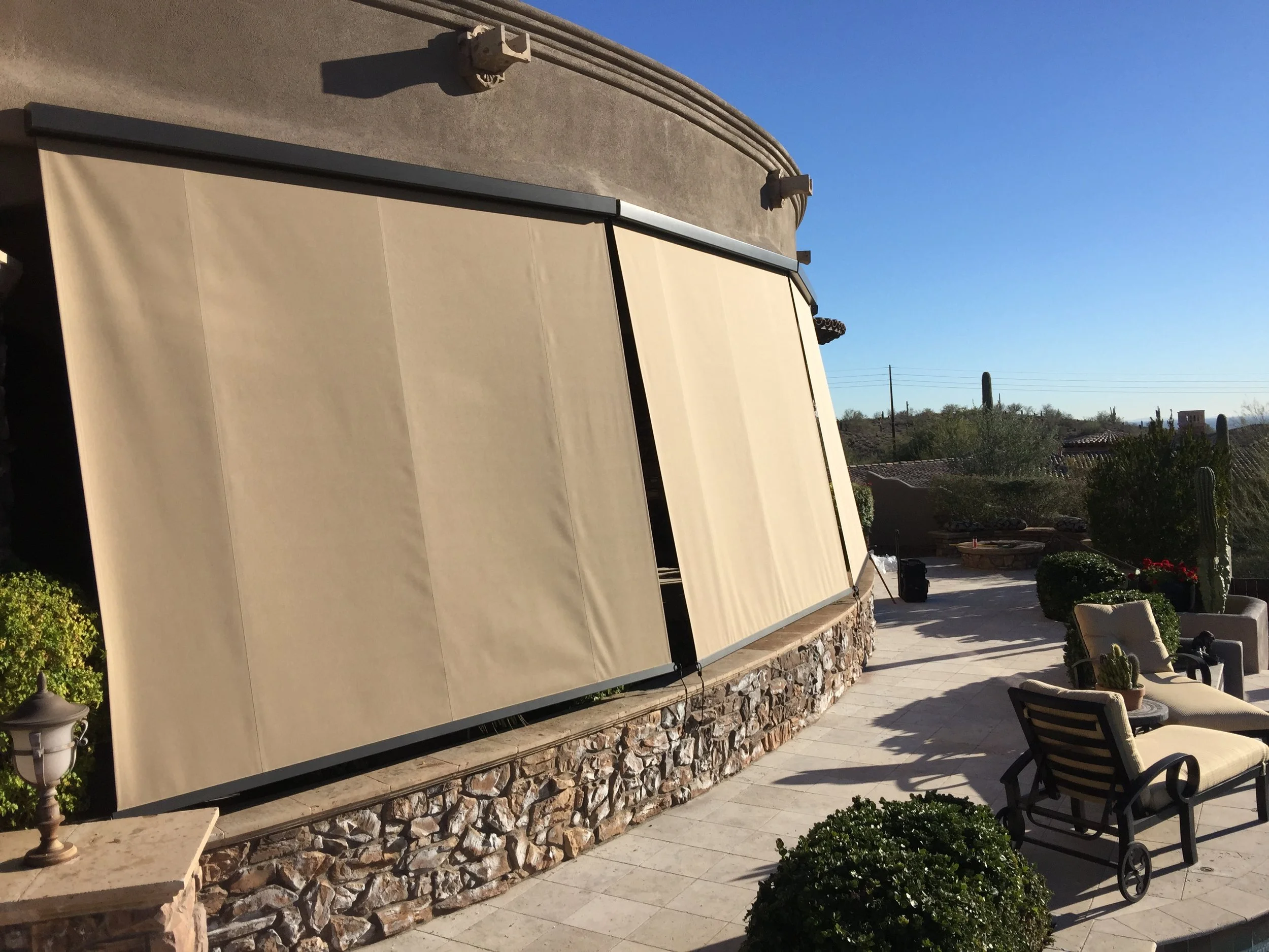 Patio area with beige retractable awnings, outdoor chairs, potted plants, and desert vegetation in the background.