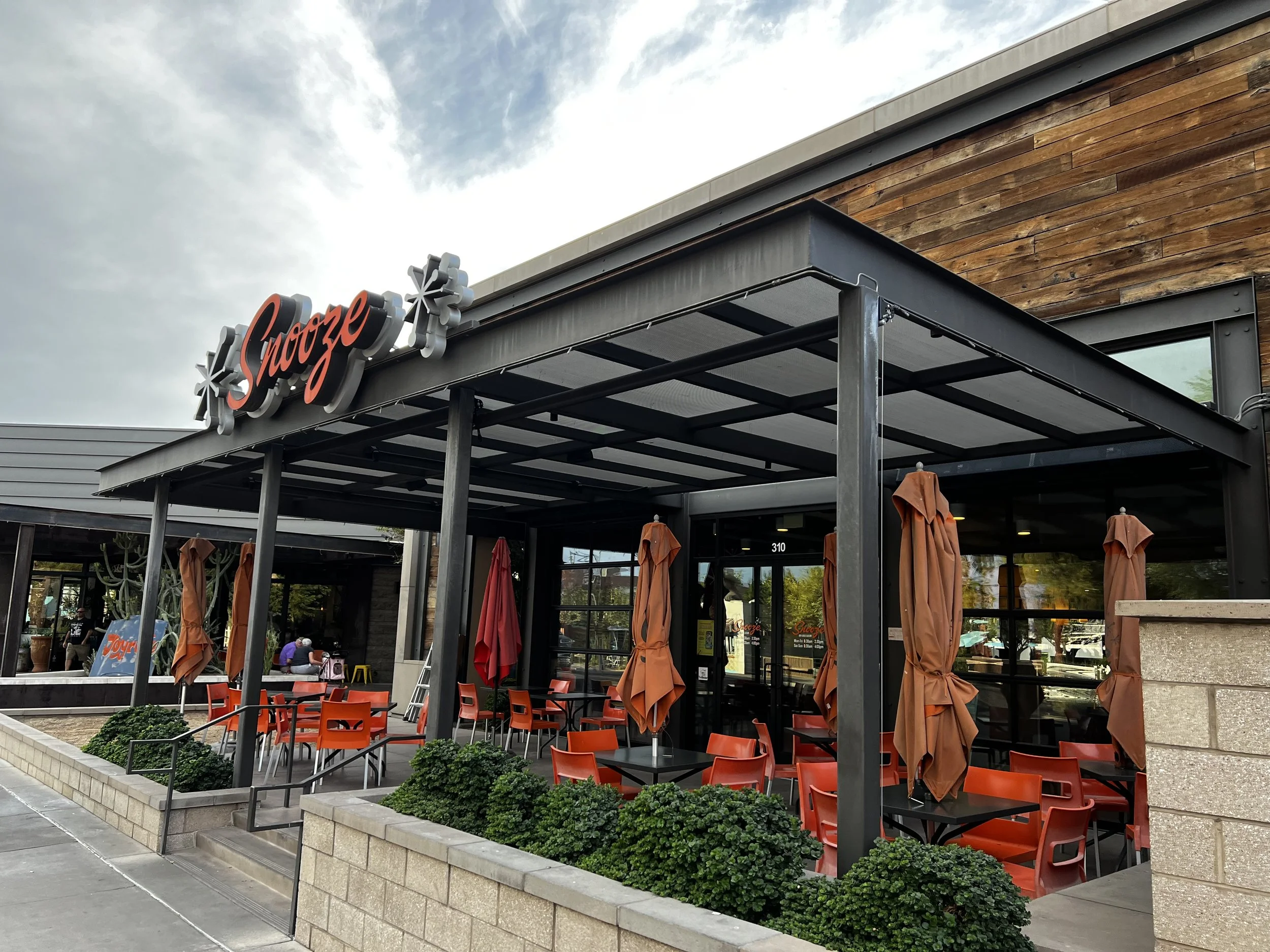 Exterior view of a restaurant with an outdoor patio featuring orange chairs, closed umbrellas, and a black metal awning. The restaurant sign reads 'Snooze' in red cursive letters.