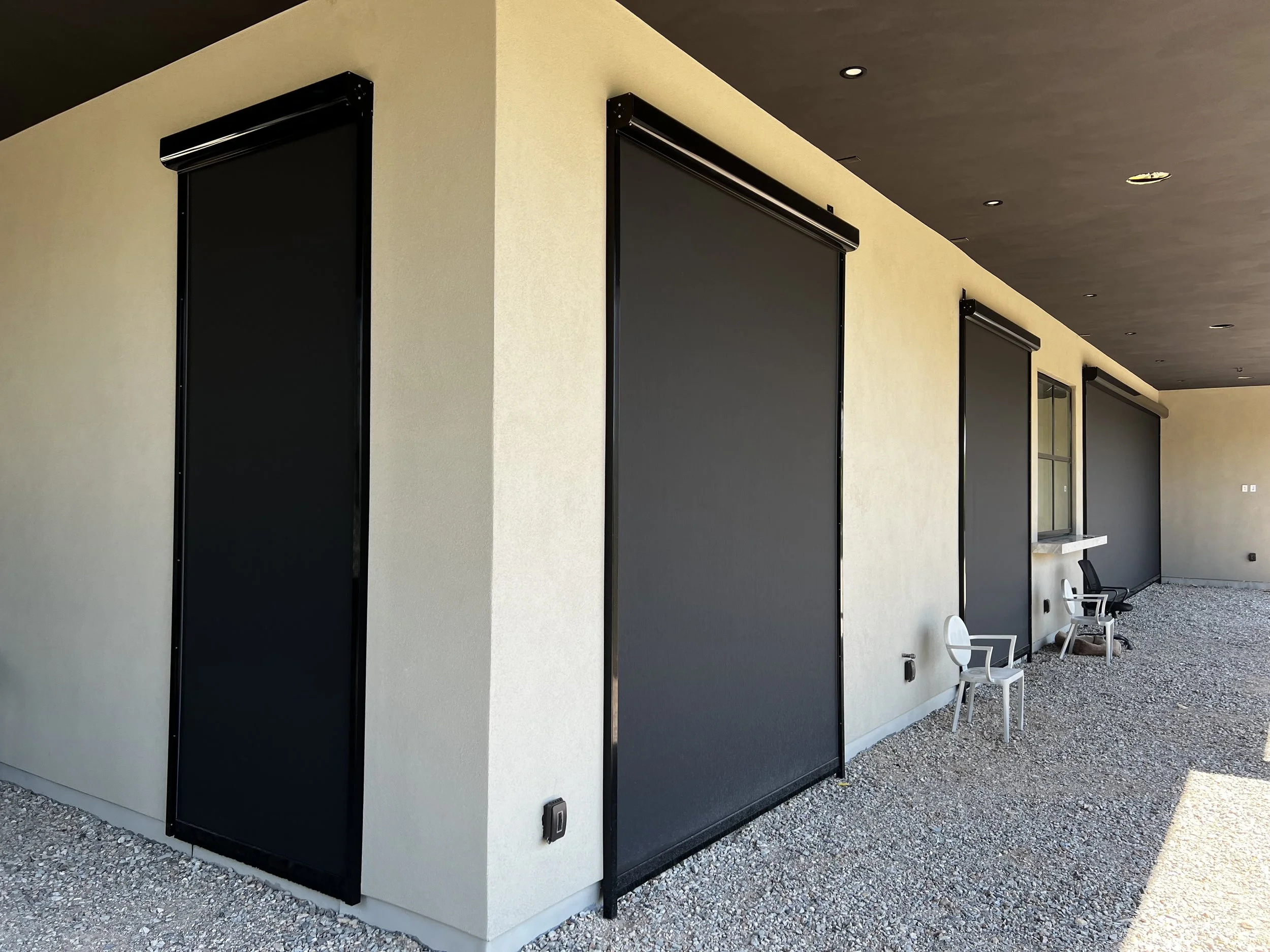 Building exterior with four closed black roller shades on beige walls, two chairs near the wall, and a small window.