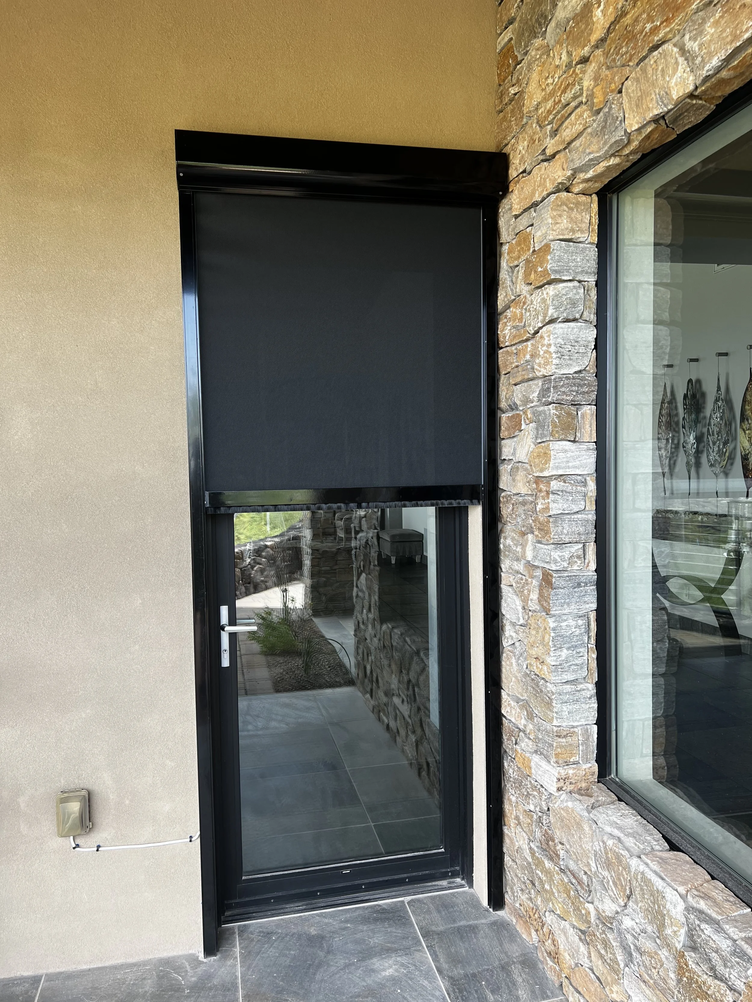 A glass door with a black retractable screen, bordered by a stone wall on the right and beige wall on the left, with a partial view of decorated interior through the glass.