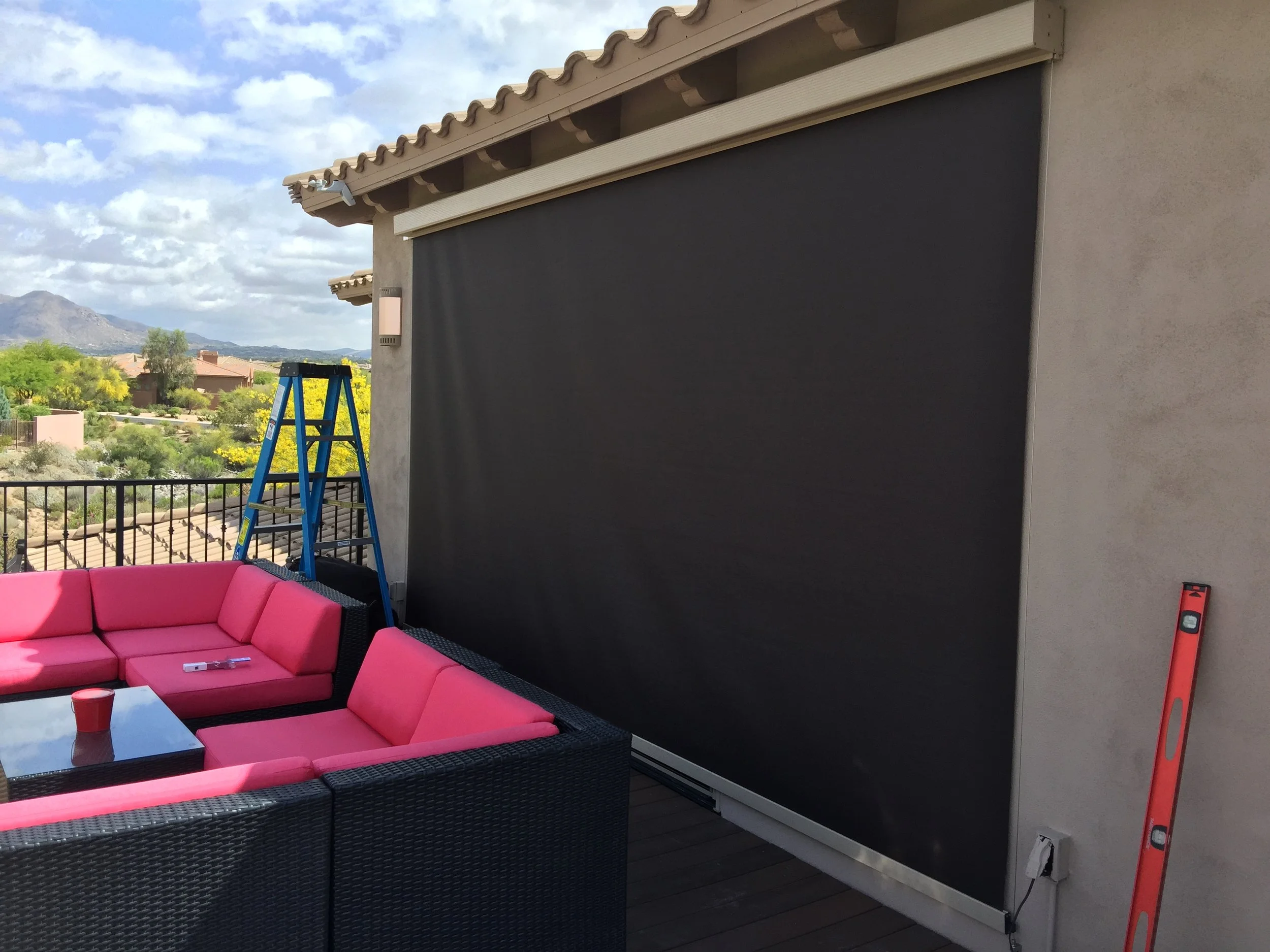 Rooftop patio scene with pink cushioned sectional sofa, a blue ladder, and a large black retractable shade on the wall, with a scenic view of houses, trees, and mountains in the background.