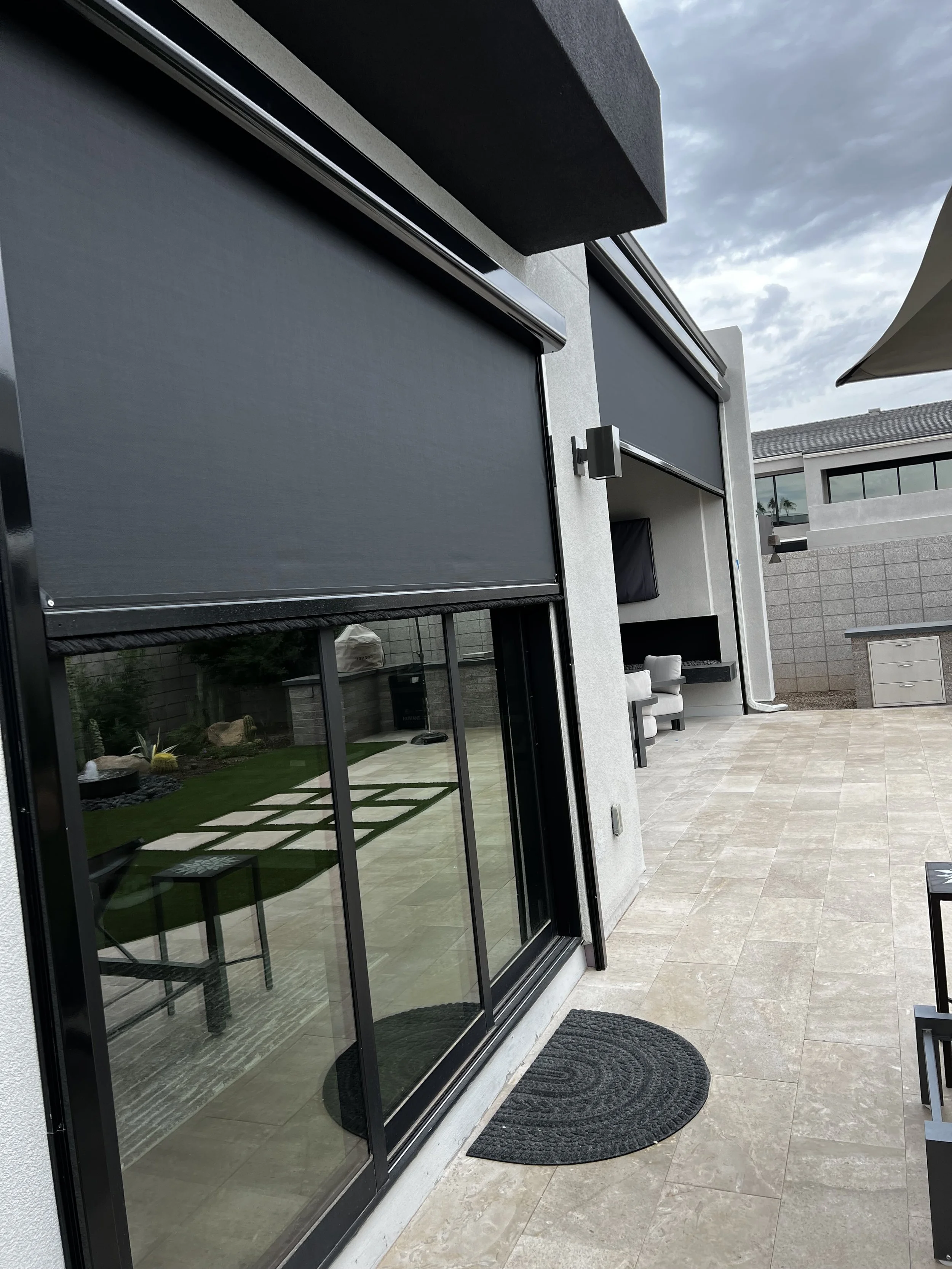 Exterior patio area with large sliding glass door, partially rolled down black shades, outdoor seating, and a brick wall enclosure.