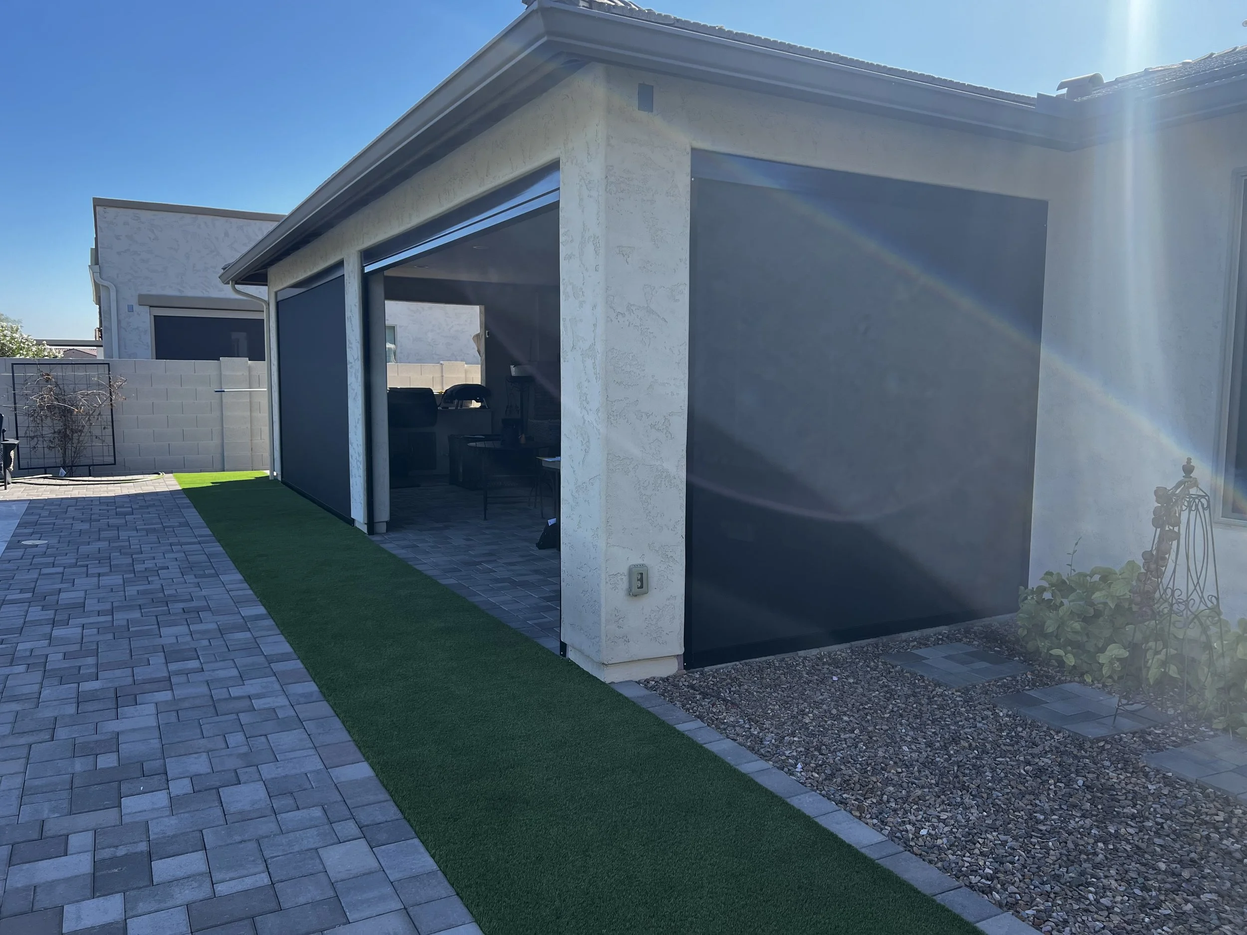 Backyard patio with paver flooring, artificial grass, and screened-in porch area under a sunny sky.
