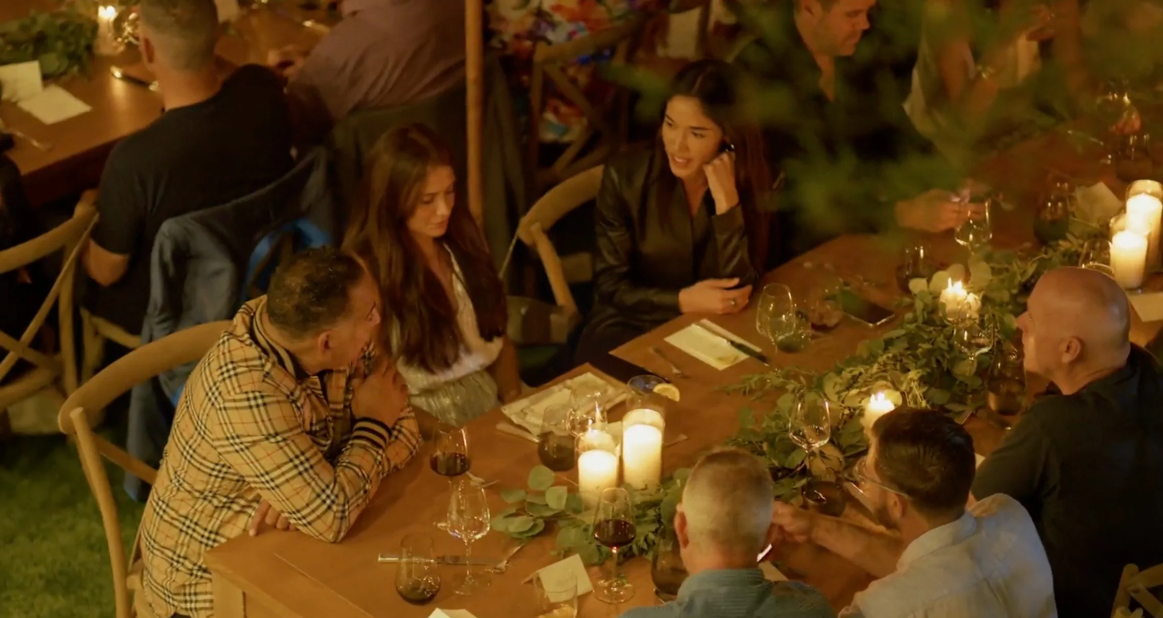 A group of people gathered around a dinner table decorated with candles and greens, socializing at an evening event.