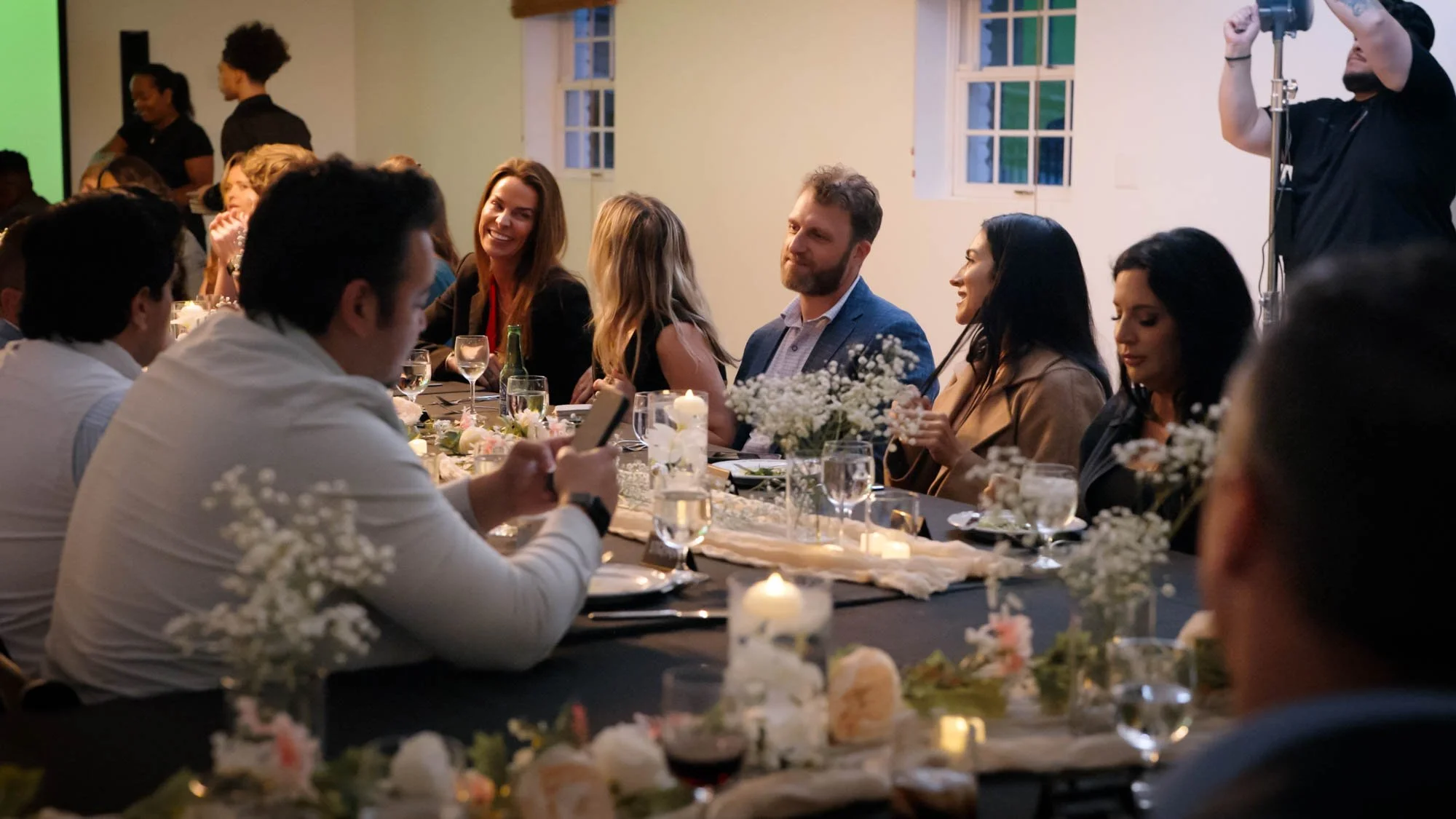 People sitting at a long decorated dinner table, smiling and talking, with floral centerpieces and candles, in a warmly lit room with windows and a projector screen in the background.