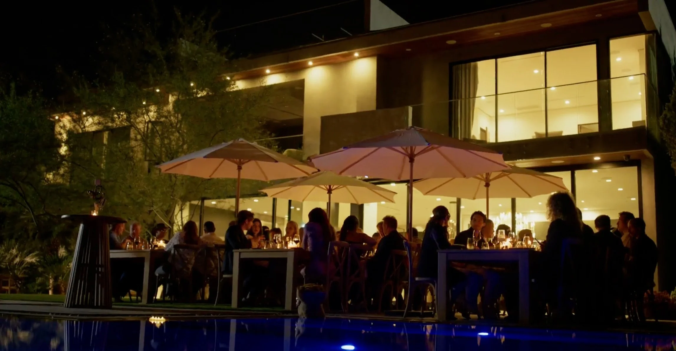 People dining outdoors at night under umbrellas at a modern house with large windows.