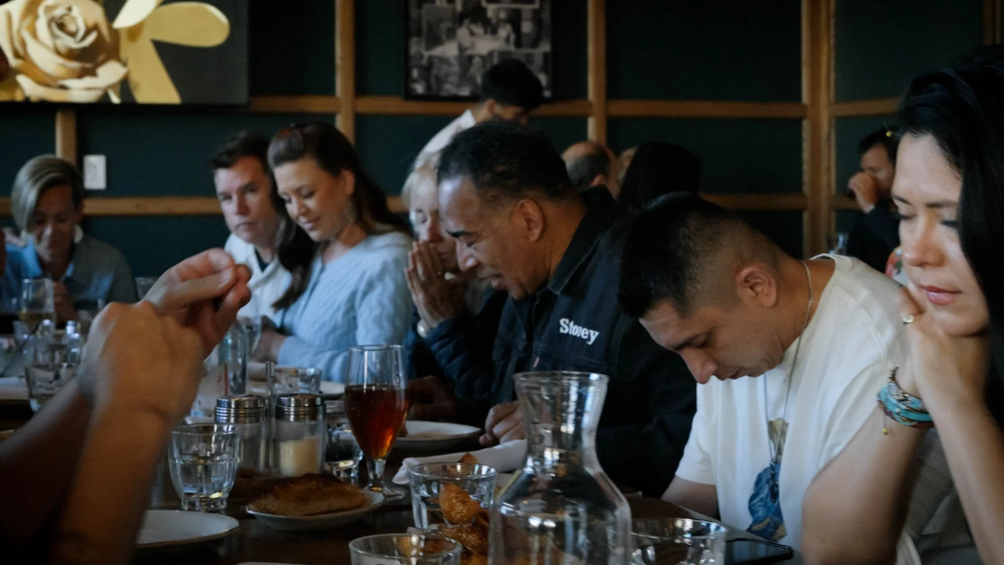 A group of people sitting at a restaurant table with their heads bowed and eyes closed, possibly praying or reflecting, with various drinks and plates of food on the table.