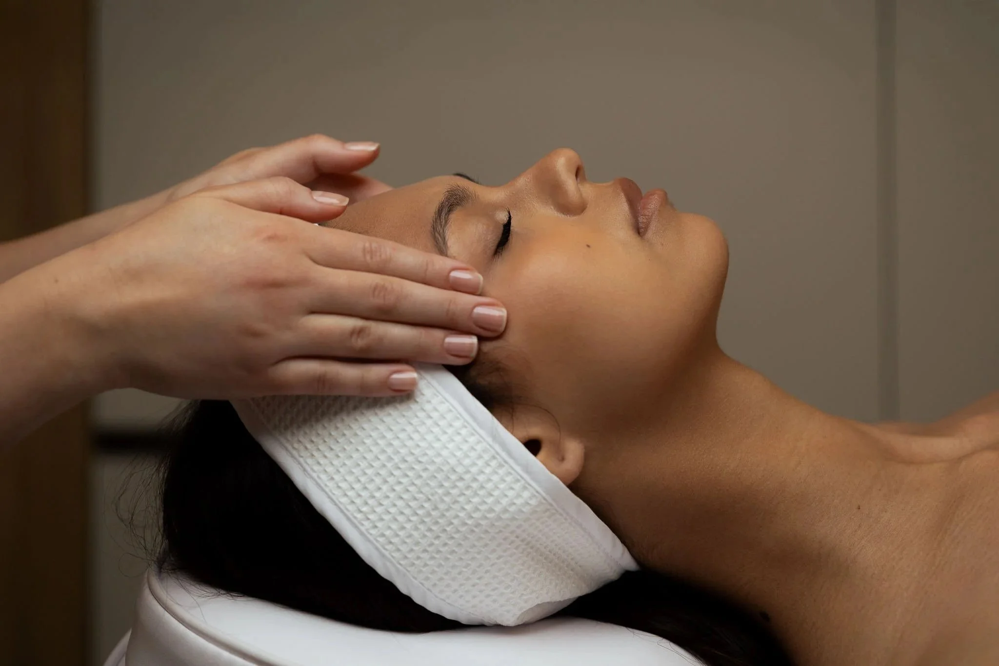 A woman laying down, her eyes closed, while a medical aesthetician rests their hands on the woman's temples.