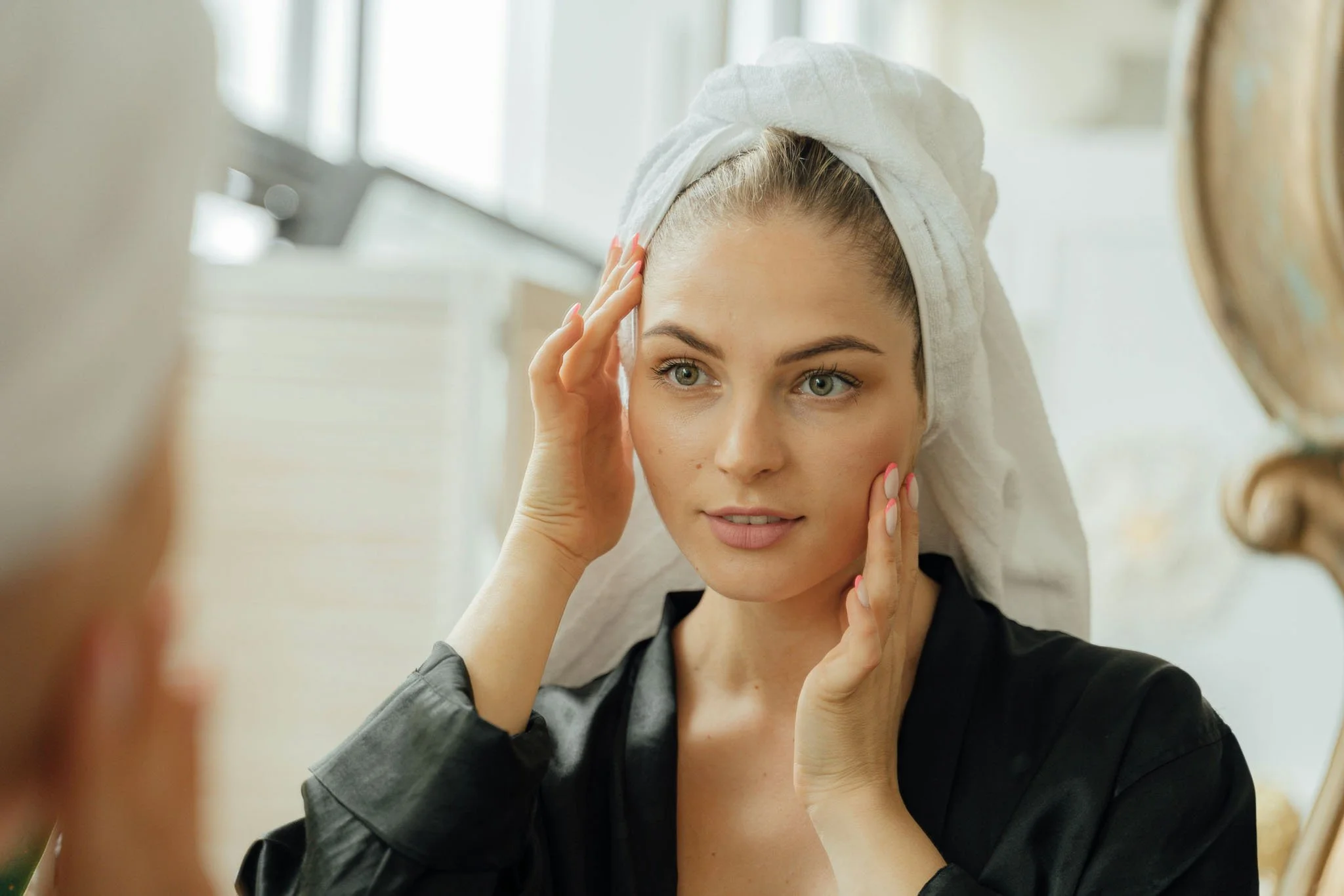 A woman with her hair in a towel and a black blouse looking at herself in the mirror while touching her face with her fingers.