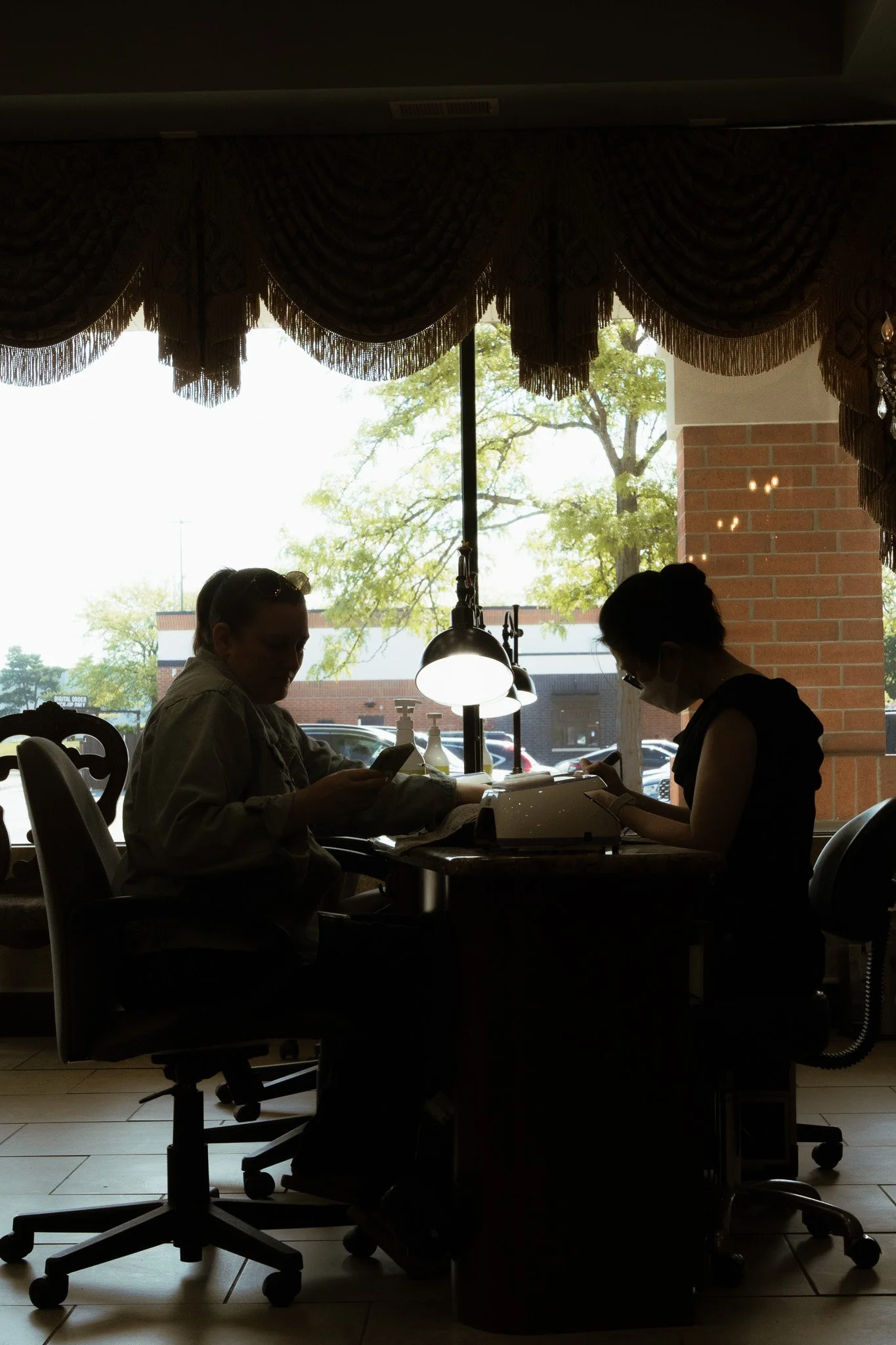 Silhouettes of two women sitting at a table in a restaurant or cafe, using their phones. Large window behind them shows trees and parked cars outside.