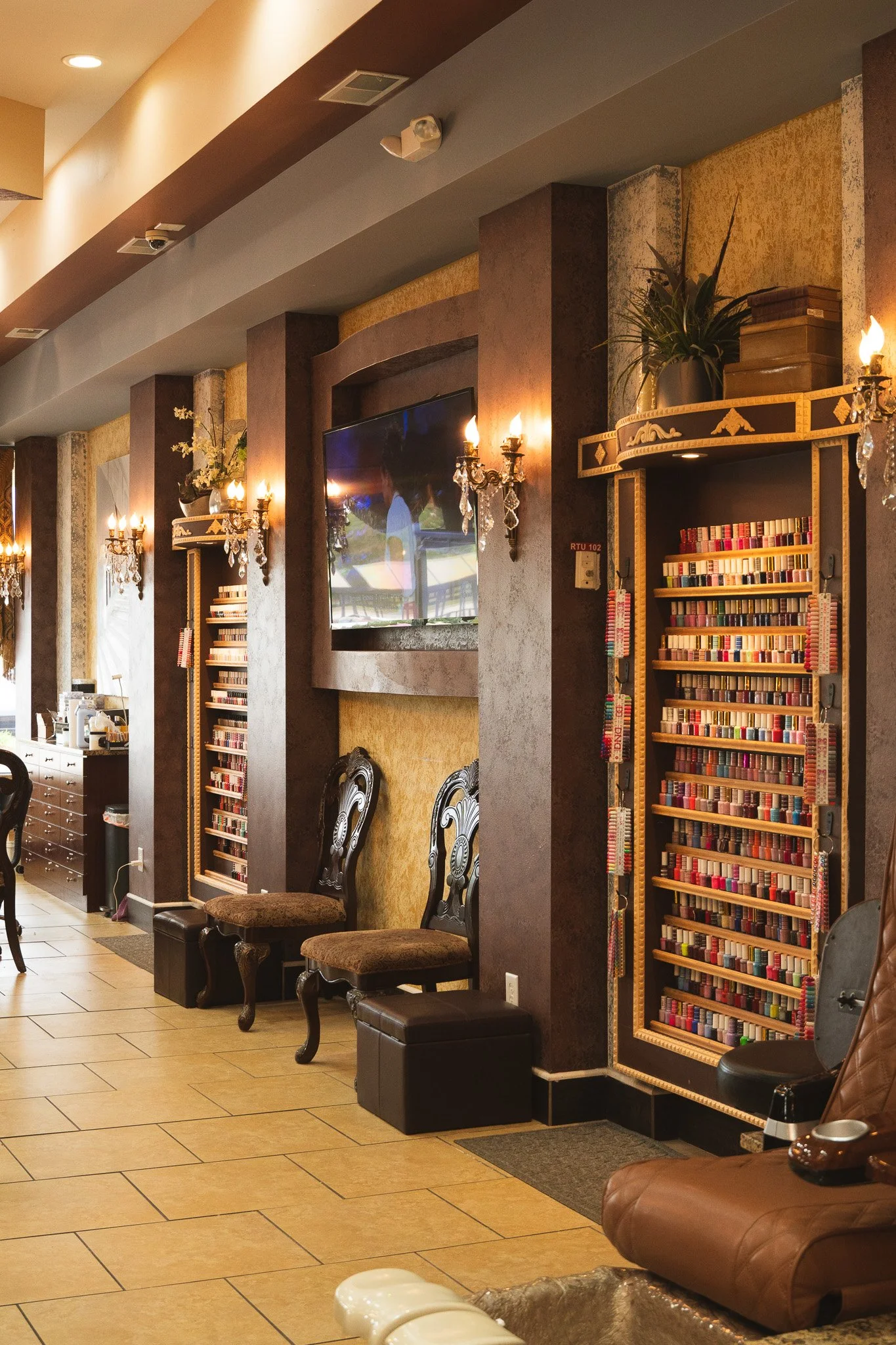 Interior of a cozy lounge or waiting area with decorative wall sconces, a flat-screen TV, two ornate chairs, a bookshelf filled with colorful books, and a leather armchair.