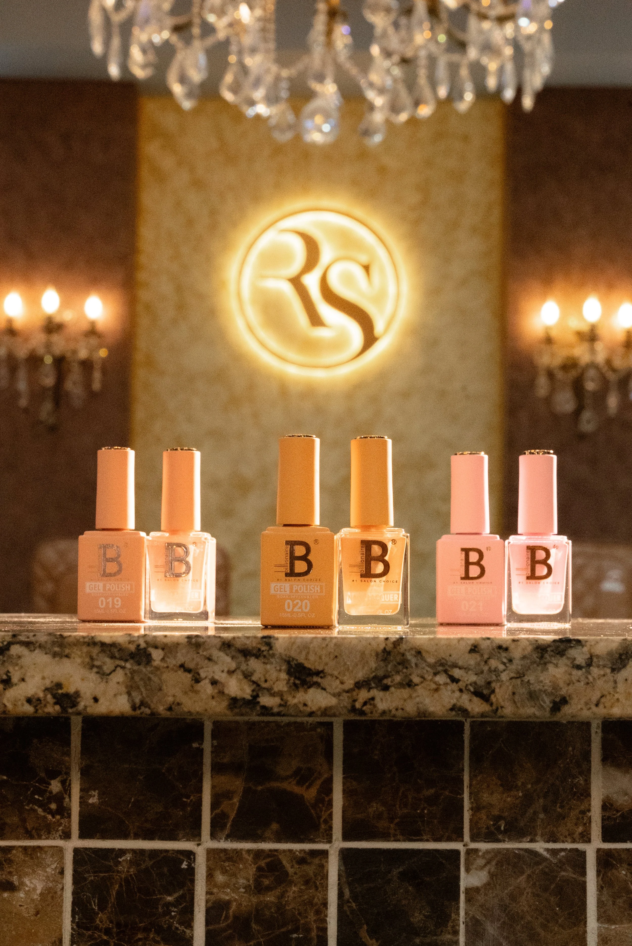 Six bottles of pink and clear gel nail polish arranged on a granite countertop, with a wall featuring a logo and chandelier in the background.