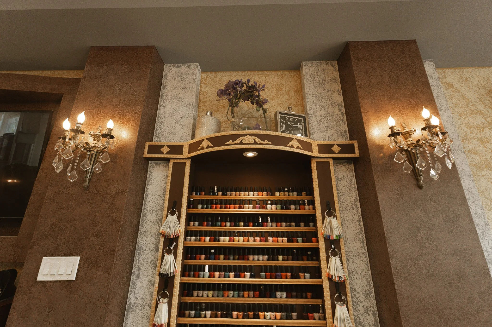 Interior of a room with a wall-mounted display of nail polish bottles, decorative wall sconces, and a shelf with decorative items including a clock, flowers, and jars.
