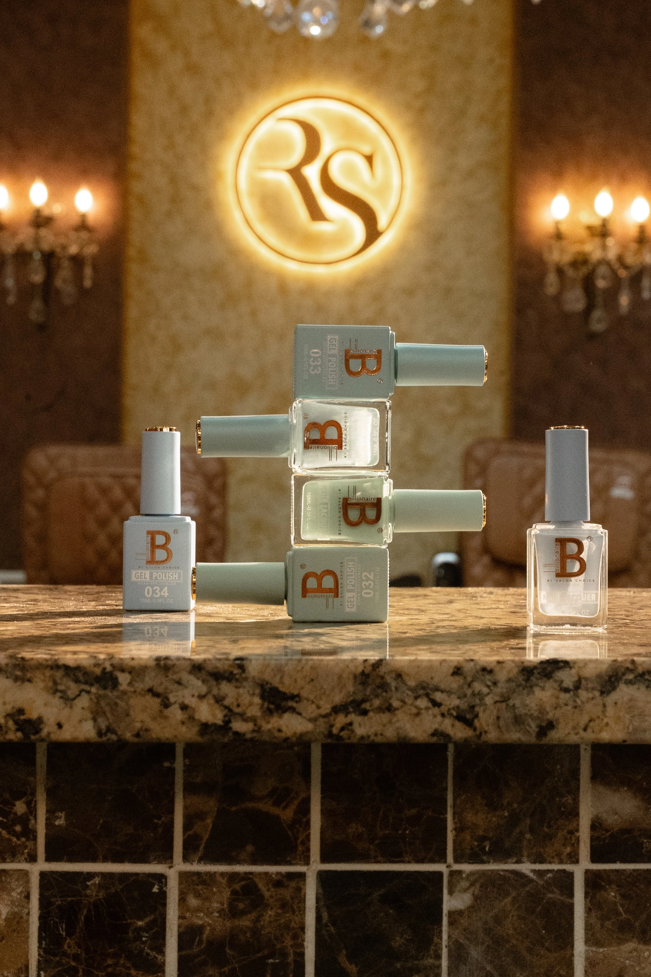 A display of several bottles of gel nail polish on a marble countertop, with a warm, elegant background featuring a circular illuminated logo with the letters 'RS' and wall sconces.
