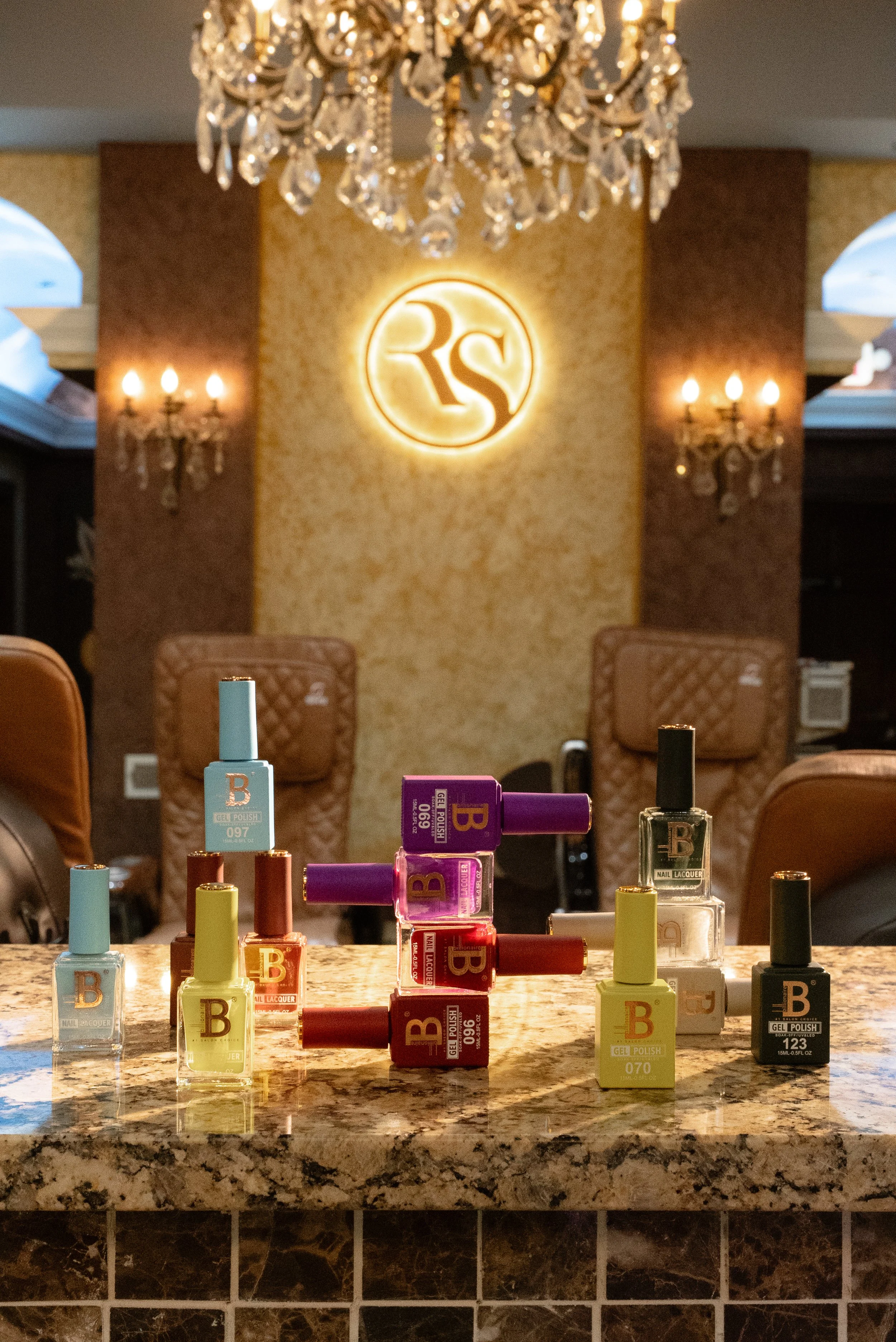 Various bottles of nail polish on a marble countertop in front of a reception area with a chandelier and illuminated RS sign on the wall.